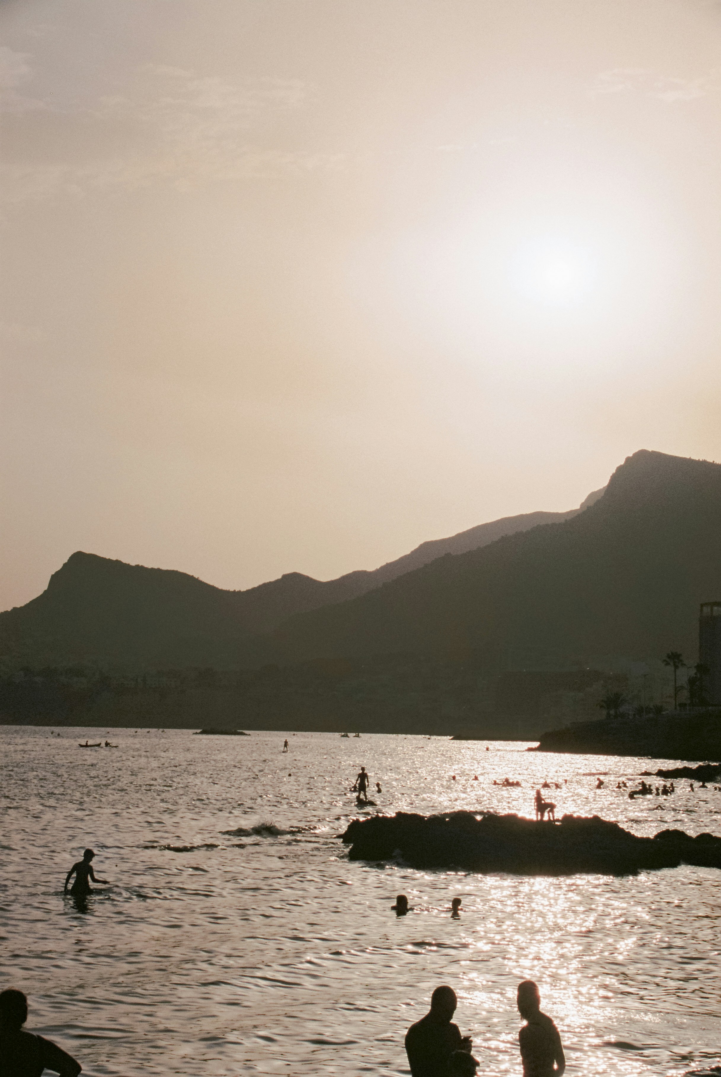 Sun-drenched coastline with distant mountains and a reflective sea, silhouettes of people enjoying the water at the shore. A soft, warm palette suggests late afternoon.
