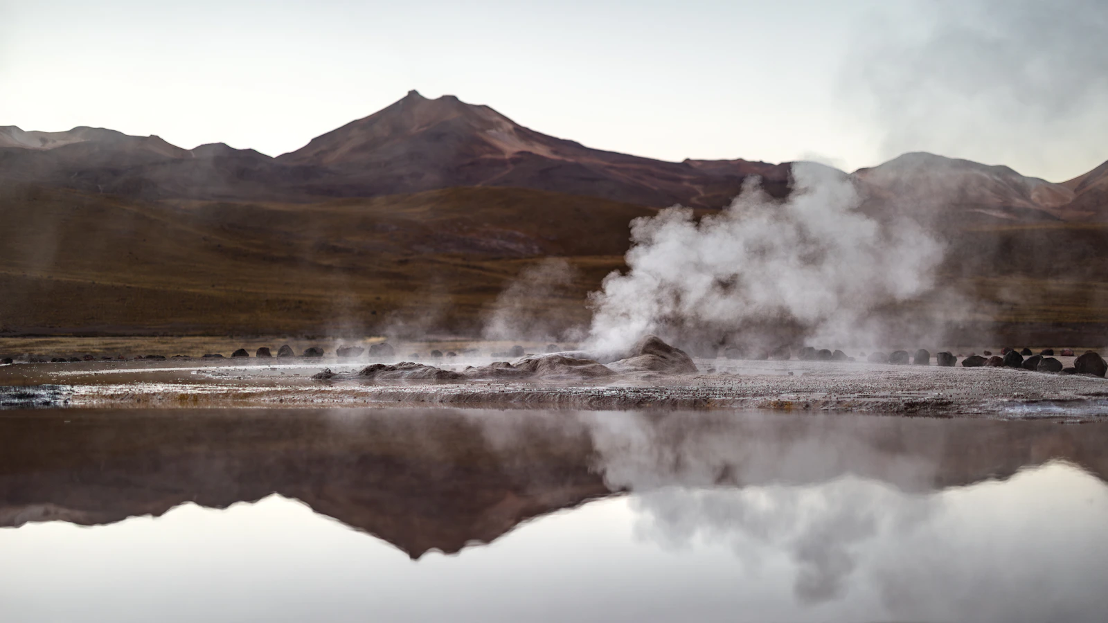 Géiser humeante reflejado en una poza con volcanes del altiplano al fondo