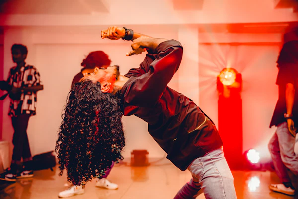 Un danseur cambré en arrière, cheveux longs bouclés, dans un studio éclairé en rouge, entouré d'autres danseurs