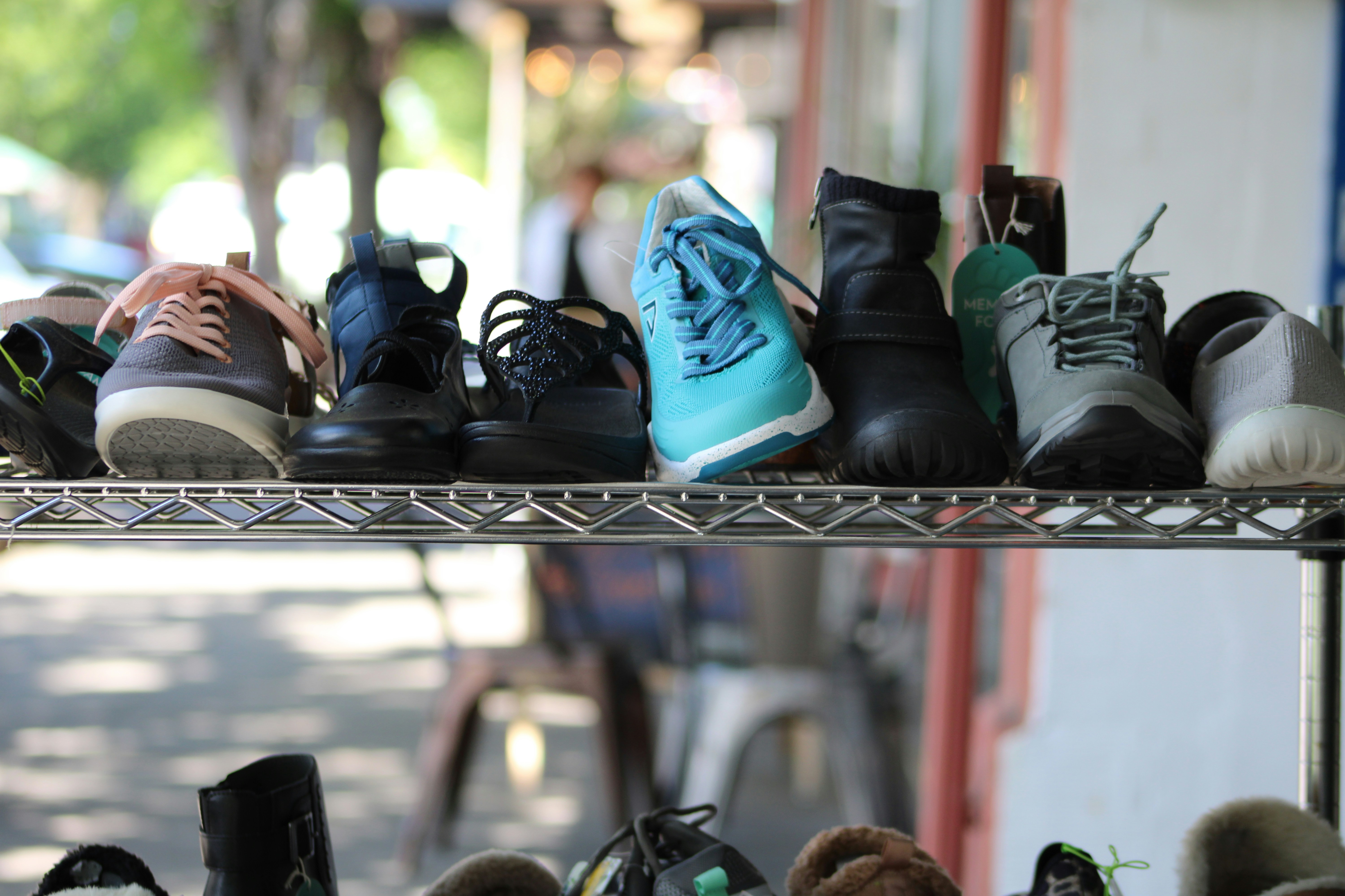 This image showcases a variety of shoes displayed on a metal rack in an outdoor setting. The shoes include different styles such as sneakers, sandals, and boots in various colors like grey, blue, black, and turquoise. The background is blurred, highlighting the shoes in the foreground, creating a sense of depth.