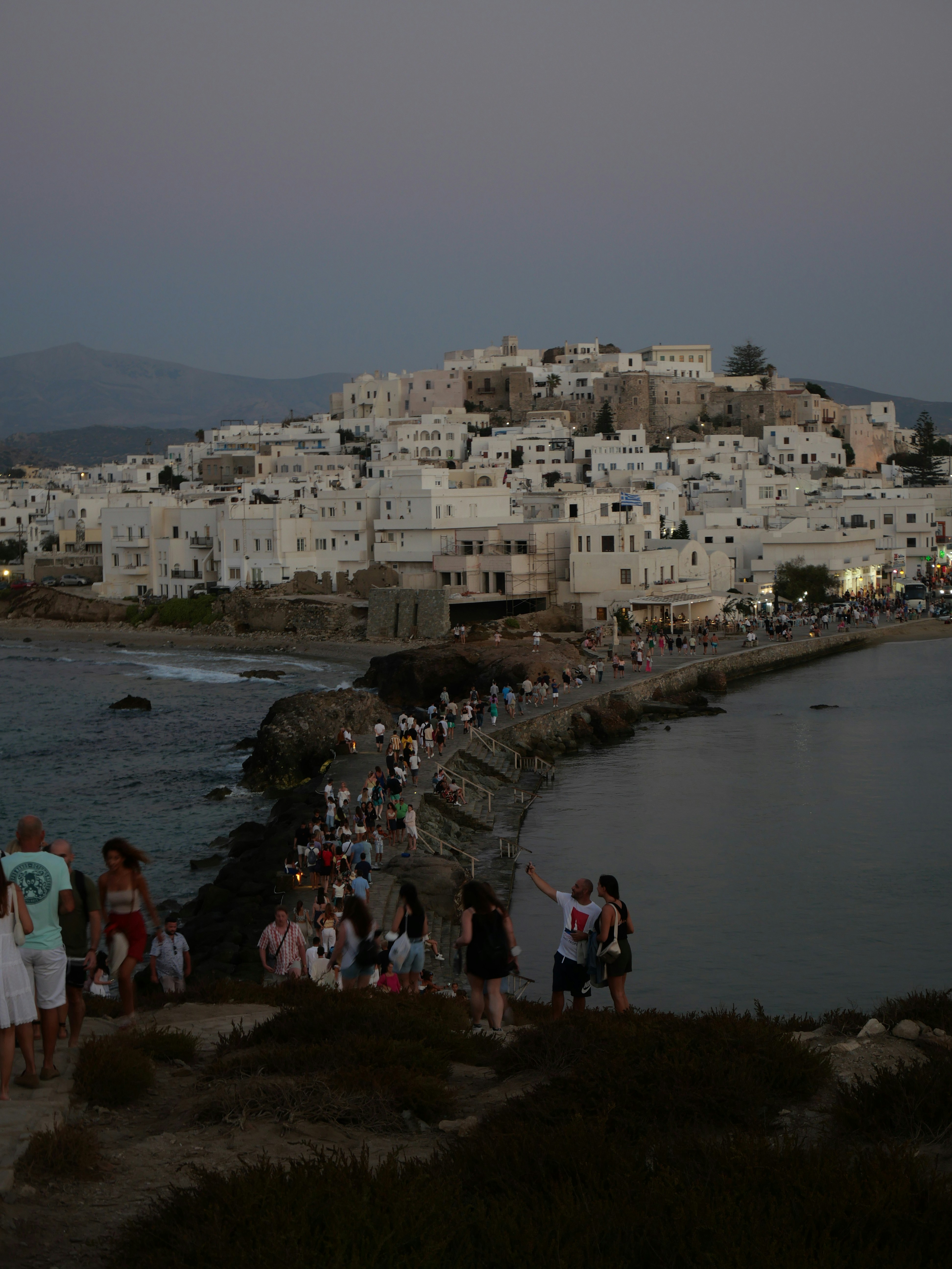 Evening photograph of a white-washed harbor village perched along a rocky promontory, with a crowded walkway along the shore. The scene captures a coastal promenade lit by dusk hues.