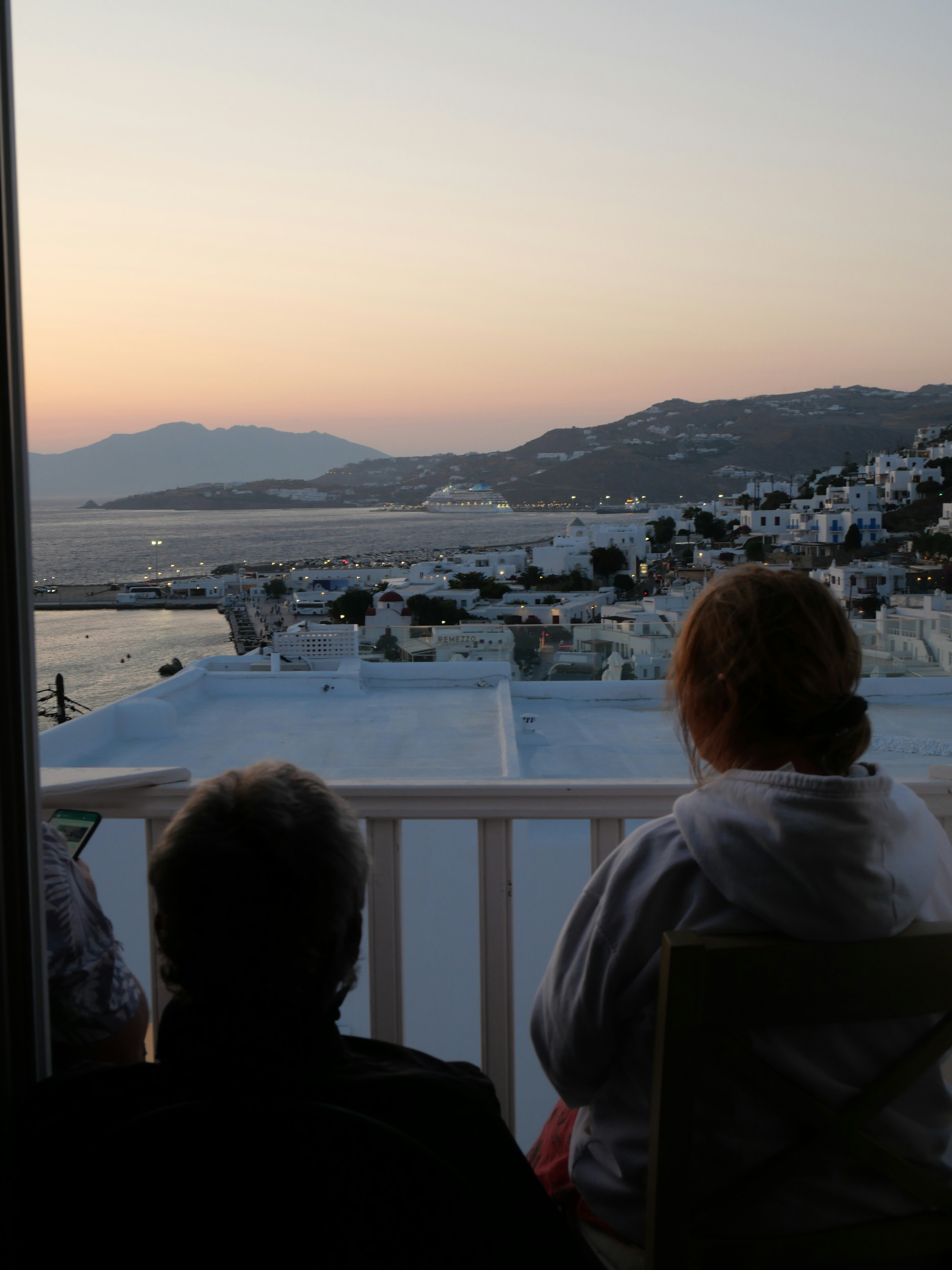 Two figures sit on a balcony, gazing toward a tranquil harbor. White-washed buildings line the coastline as a peachy sunset softens the horizon.