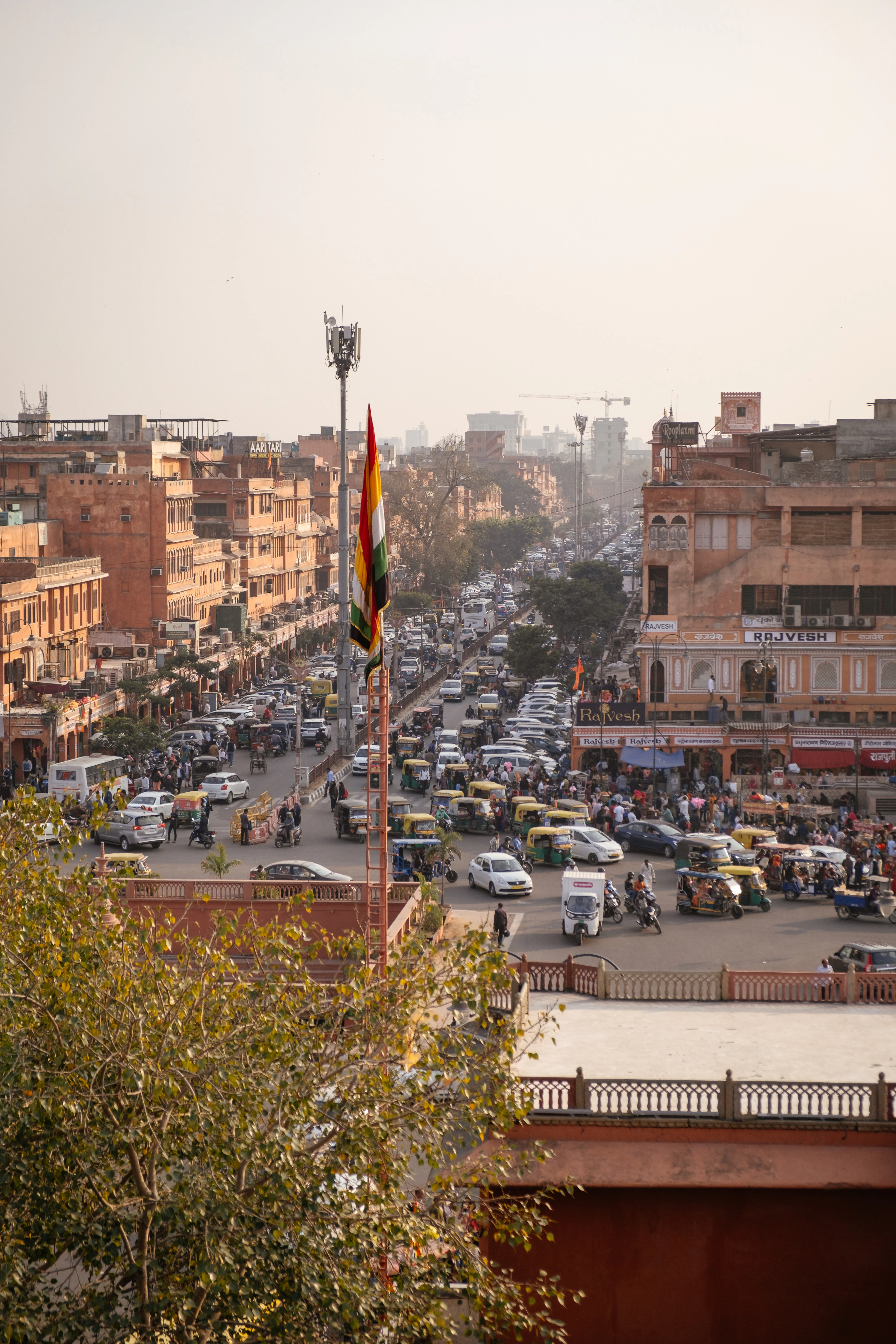 A bazaar street inside Jaipur's walled Pink City seen from above in hazy afternoon light, arcaded terracotta-pink shopfronts running down both sides and the road below packed with auto-rickshaws, scooters and shoppers