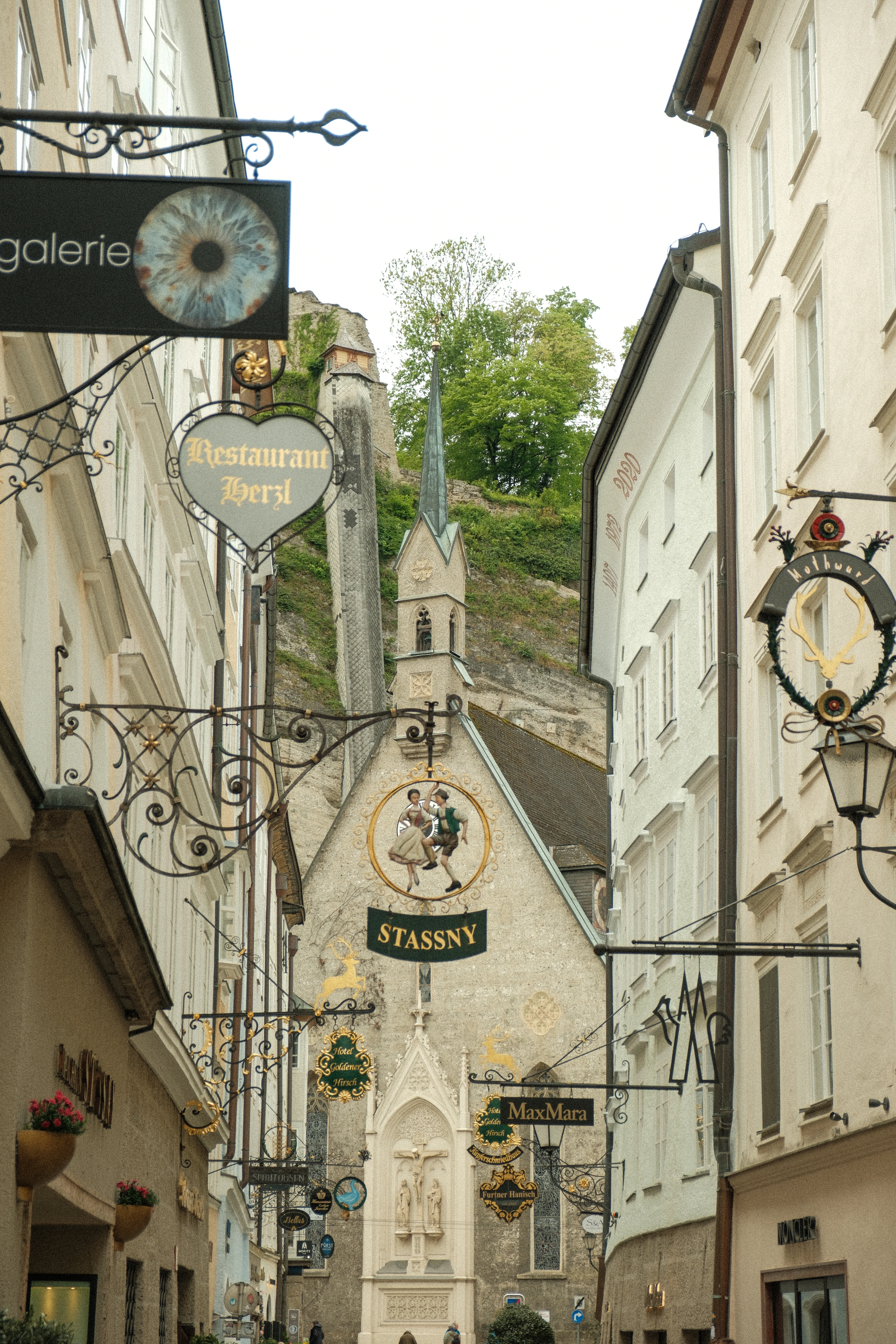 Ornate wrought-iron shop signs line a narrow old-town Salzburg street leading to a church spire.