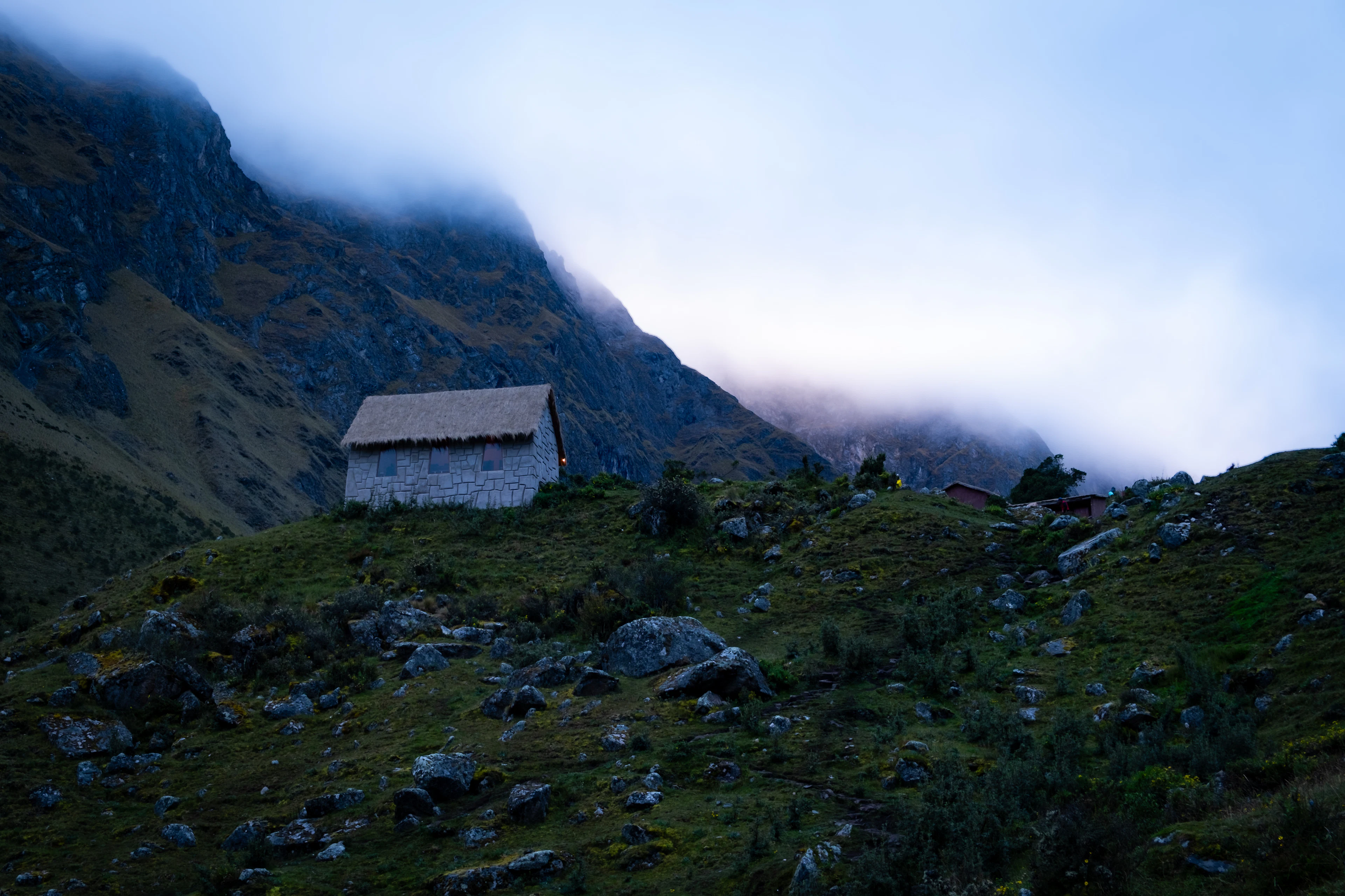 A stone hut with a thatched roof on a green slope beneath cloud-wrapped mountains, Salkantay trail, Peru
