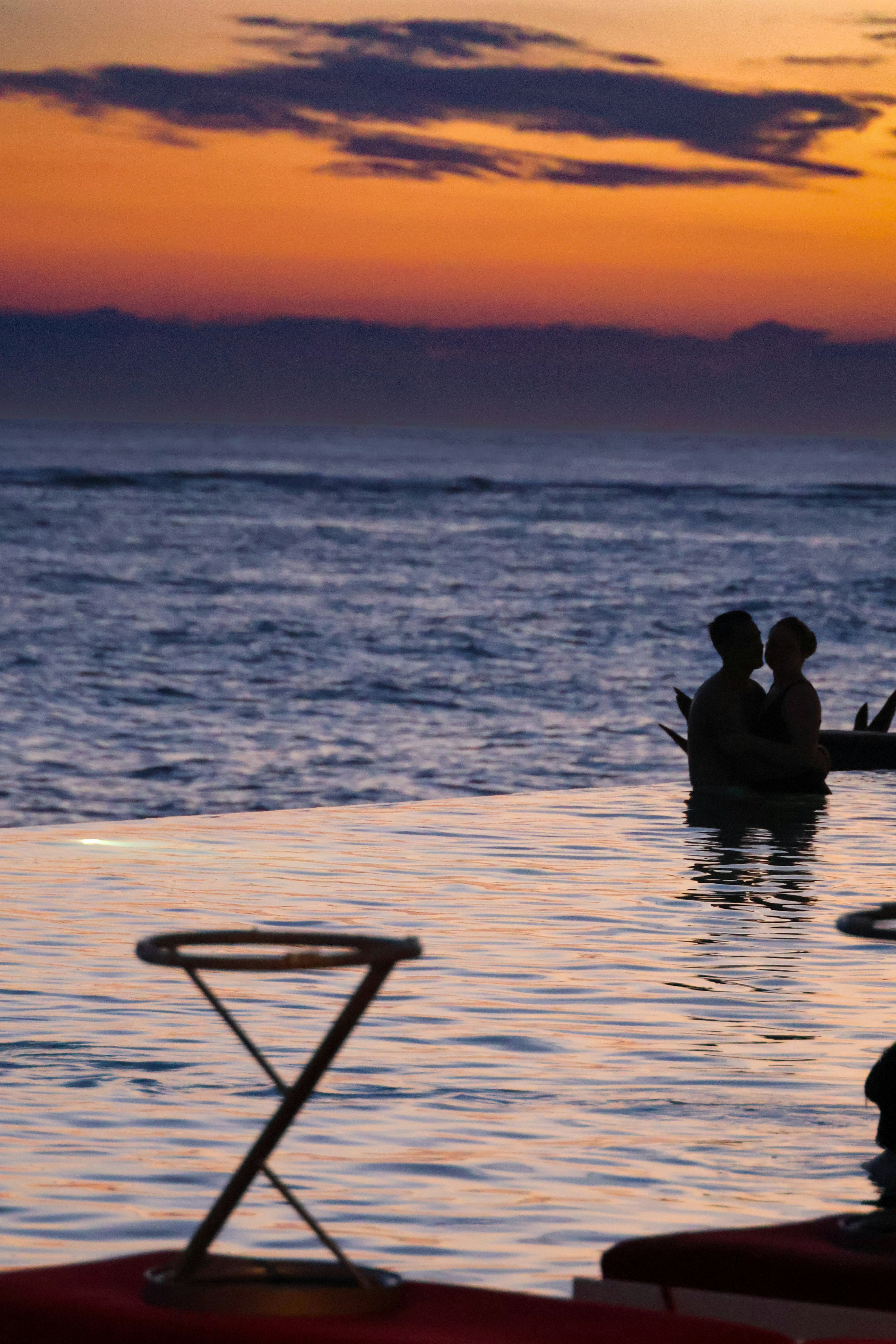 Couple silhouetted at infinity pool overlooking ocean at sunset, dramatic orange sky, deep shadows