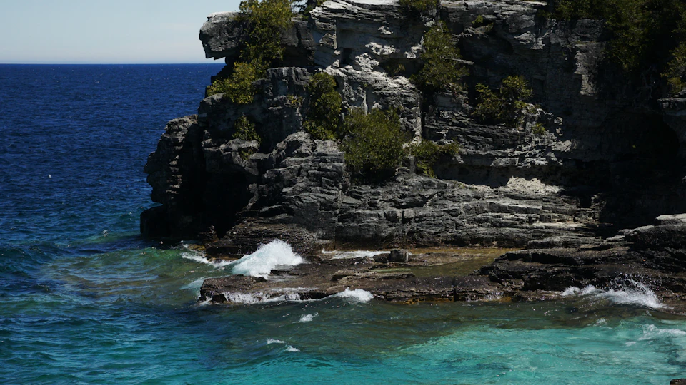 The image depicts a rocky cliff overlooking a deep blue ocean. The cliff is covered in green vegetation and features several small trees scattered across its surface, adding to the natural beauty of the scene. The water below the cliff is a vibrant shade of blue-green, with white waves crashing against the rocks. The perspective of the photo is from the shoreline, looking out towards the ocean, creating a sense of depth and vastness in the image.
