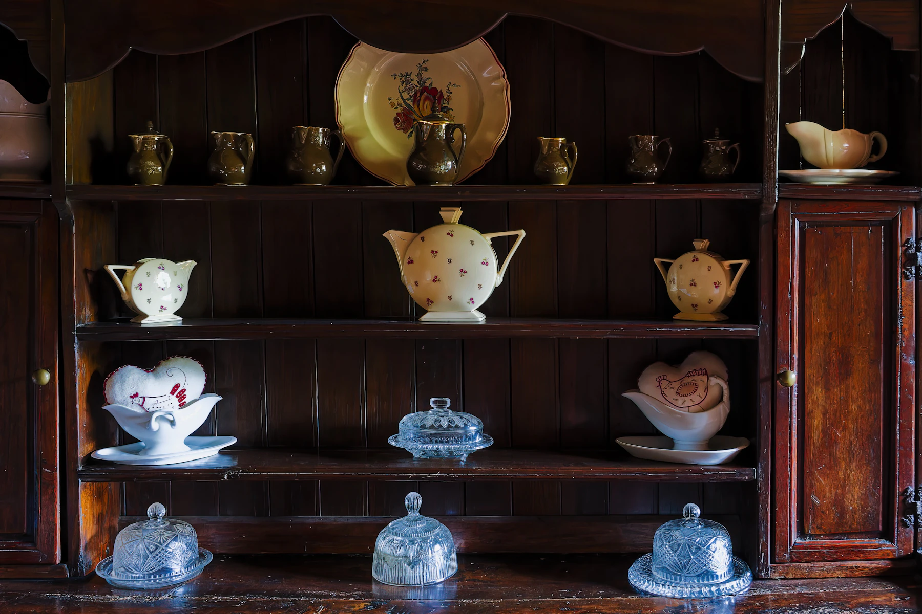 A dark wood dresser with a rose-sprigged teapot on the middle shelf, a row of small jugs above and covered glass dishes below