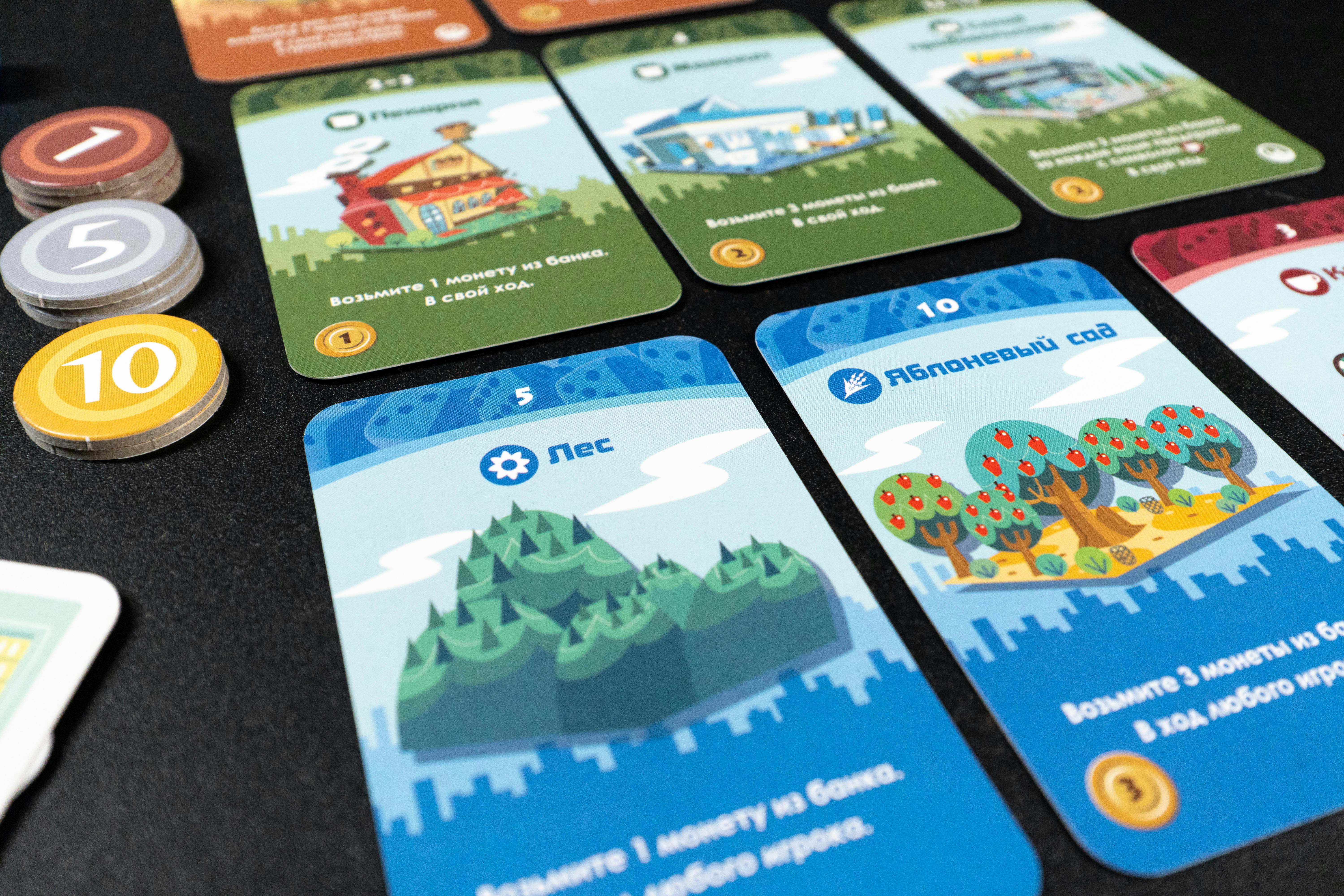 A top-down view of randomly selected cards and components from Machi Koro and its expansion. The image includes various establishment cards, coins, and dice, showcasing the diverse and vibrant elements of the game. This assortment highlights the variety of strategic options and the colorful artwork that enhances the overall gaming experience.