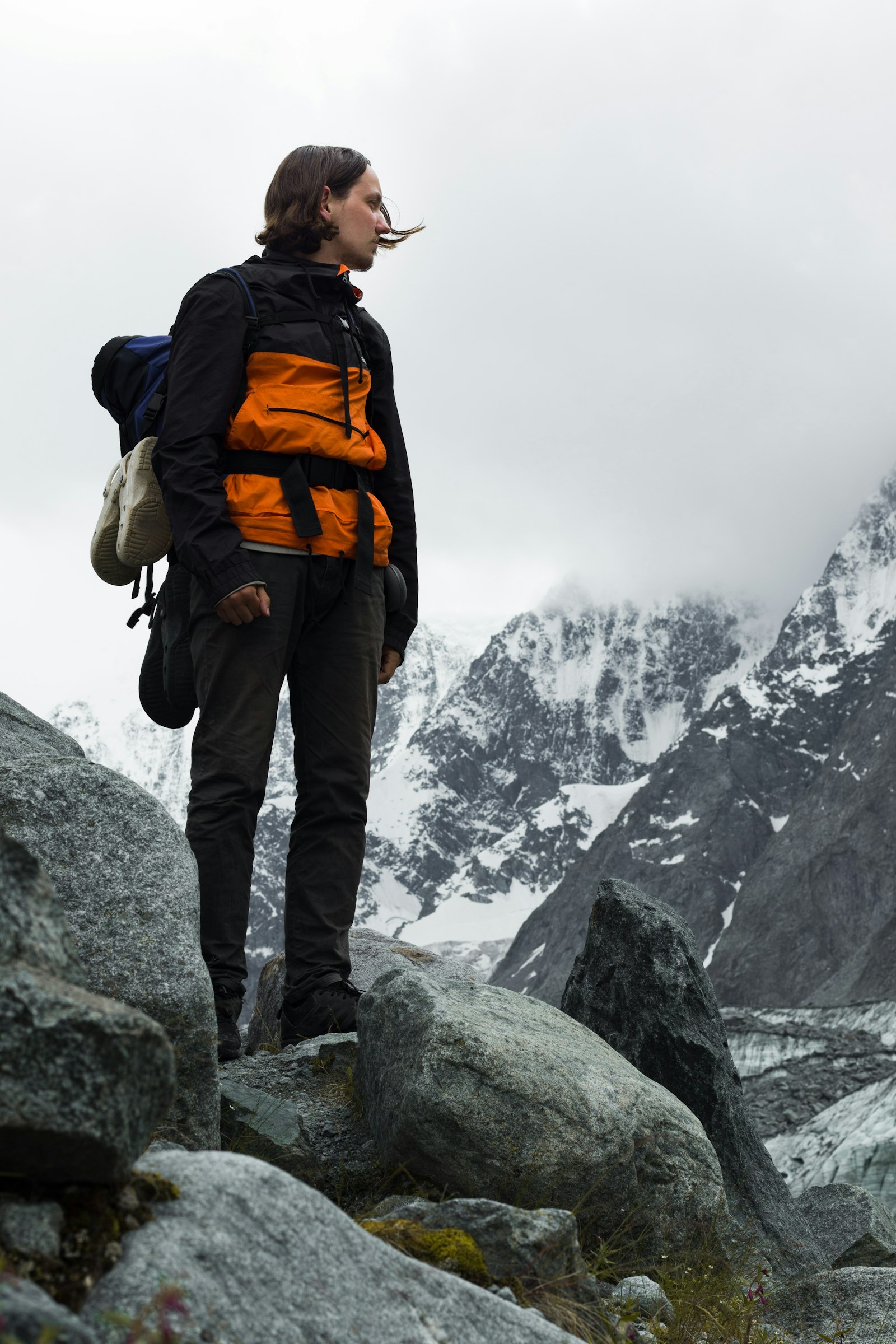 Un randonneur en veste orange et noire, debout sur des rochers face à des sommets enneigés