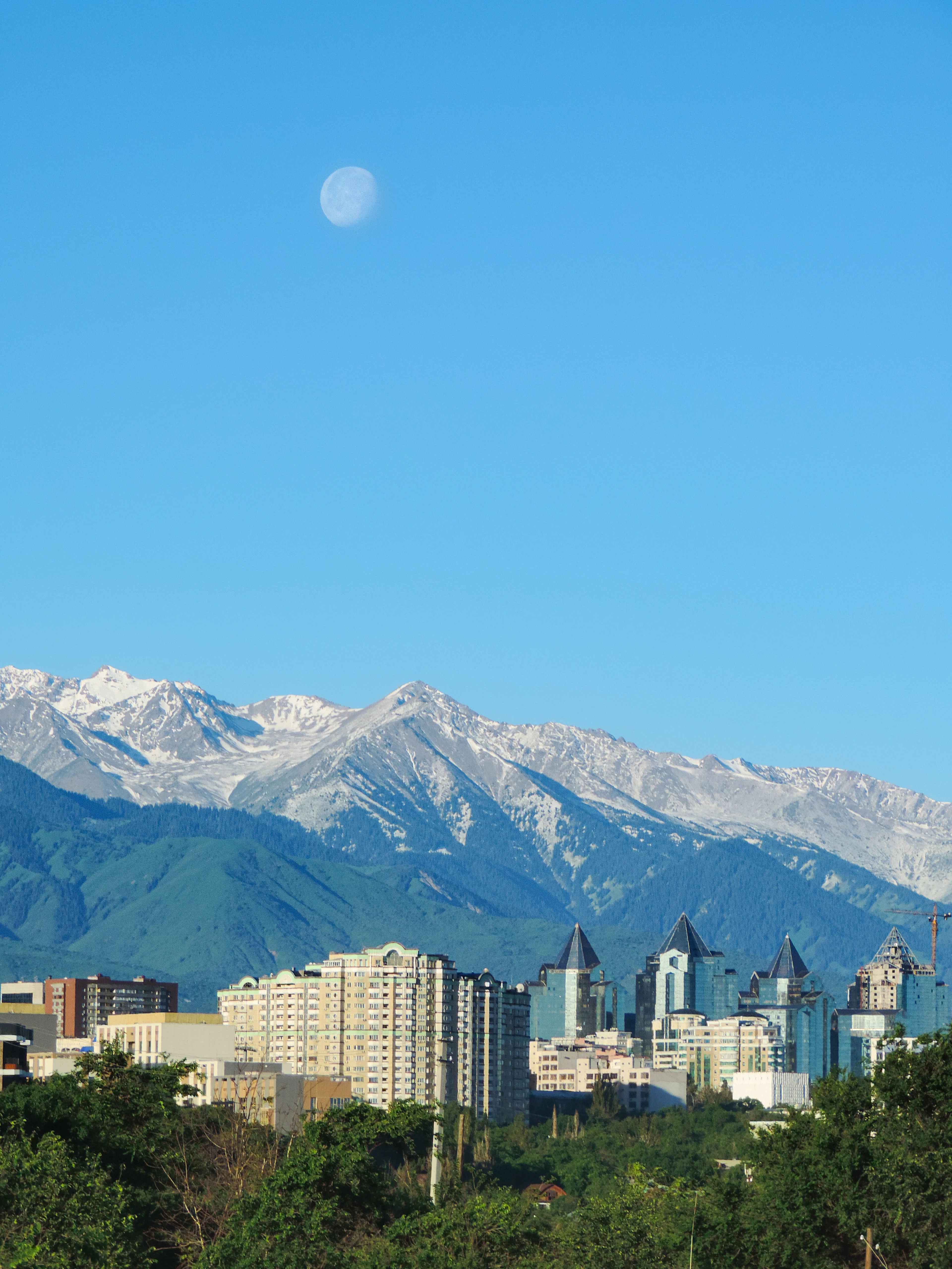A near-full moon hangs over Almaty's lit skyline at dusk, with snow-dusted mountains behind the towers.