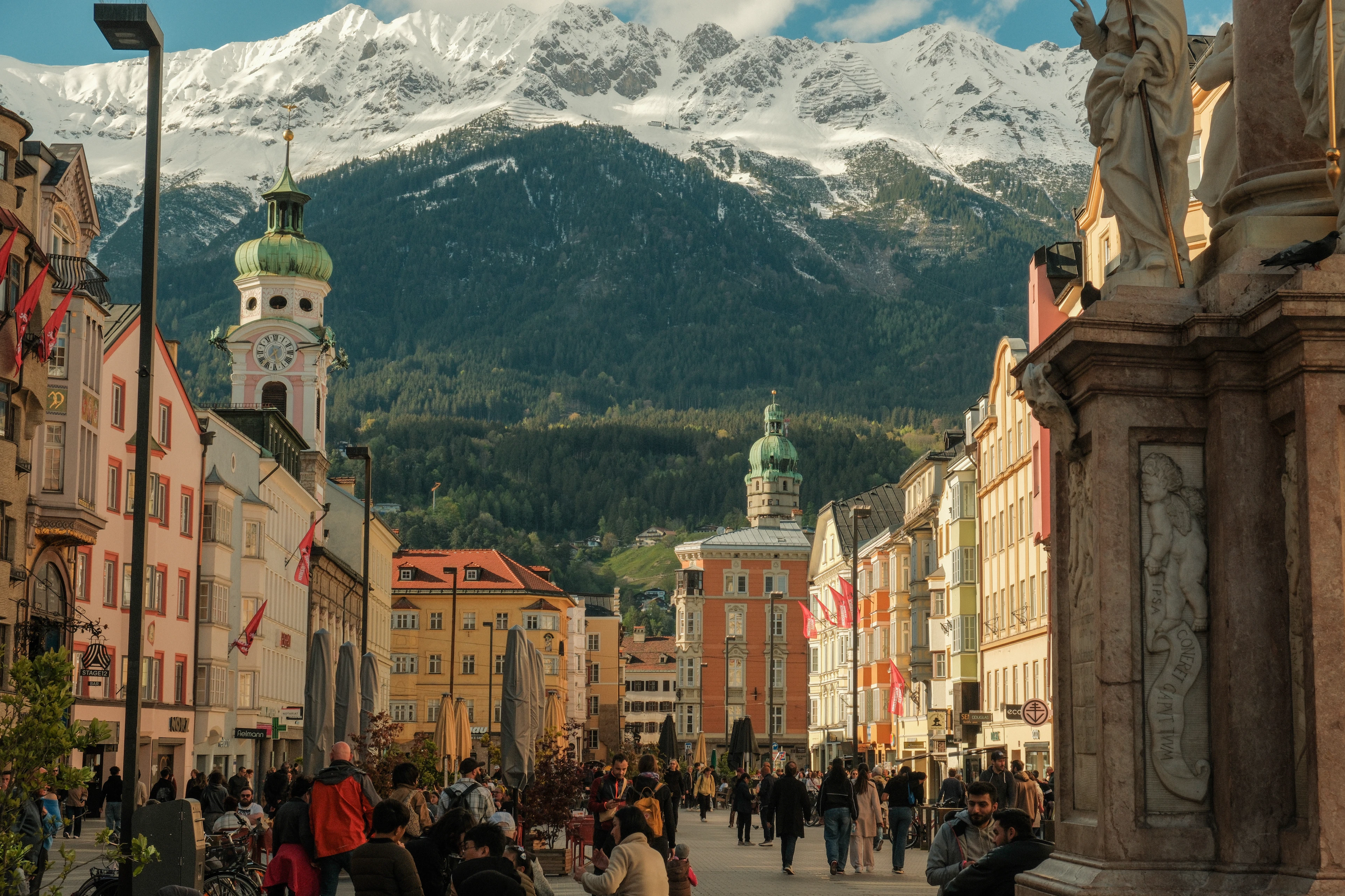 Maria-Theresien-Strasse in Innsbruck lined with pastel buildings and a statue, jagged snow-dusted peaks rising behind.
