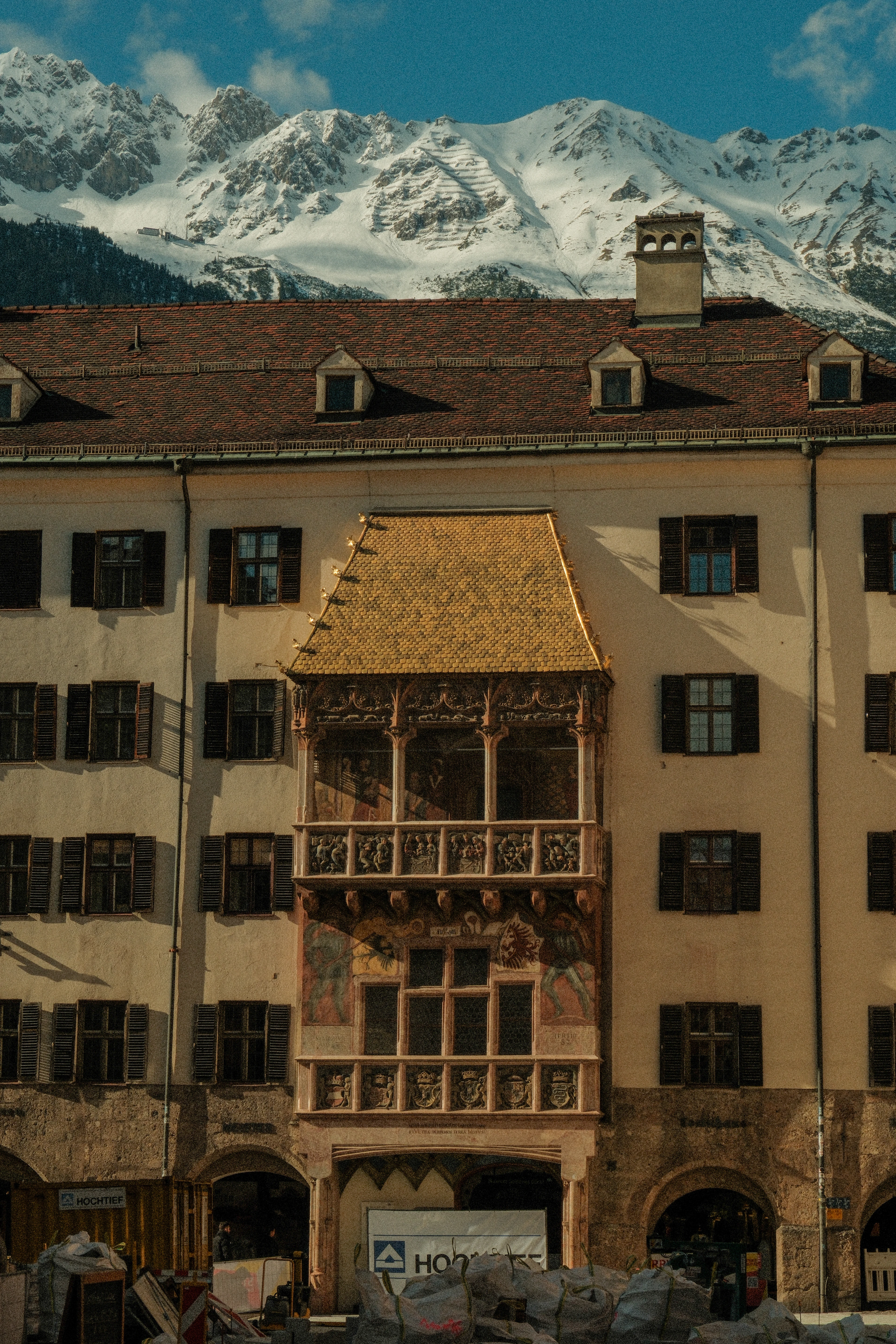 The gilded copper canopy of Innsbruck's Goldenes Dachl glowing above its frescoed oriel balcony on the pale Neuhof facade, with the snow-covered crags of the Nordkette rising directly behind the roofline under a blue sky.