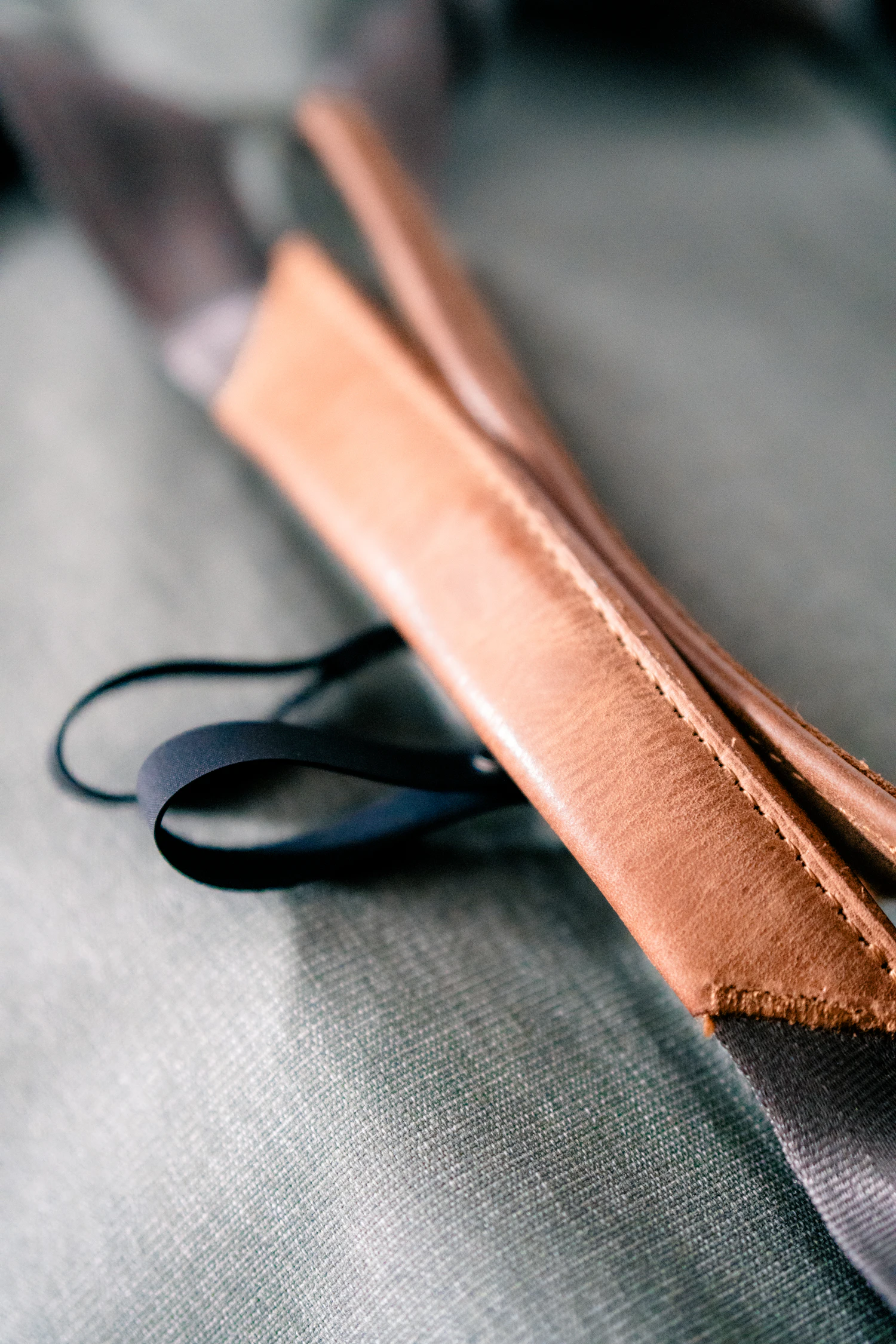 Two cut lengths of tan leather strap lying on a grey cloth beside a black webbing loop