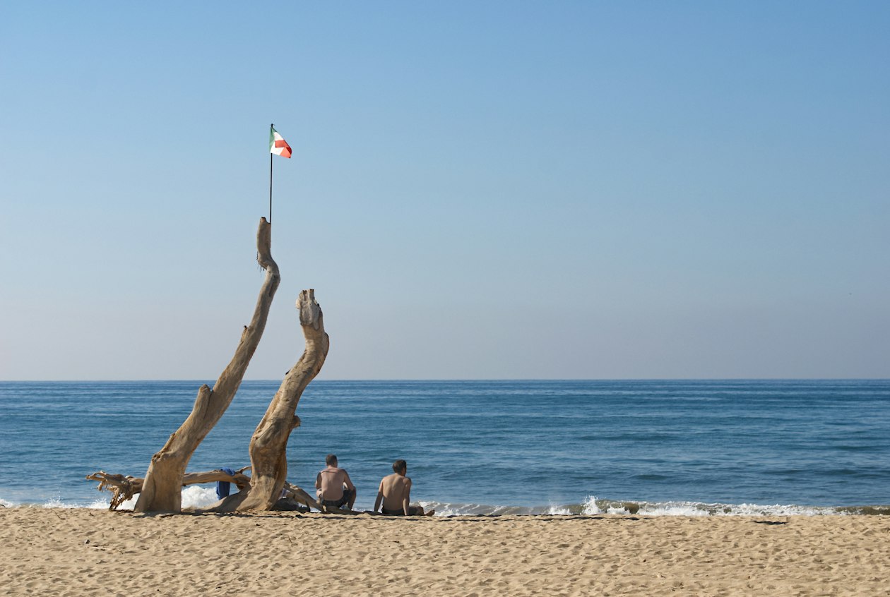 Two people sitting on the sand by driftwood under a clear blue dry-season sky on the Pacific coast