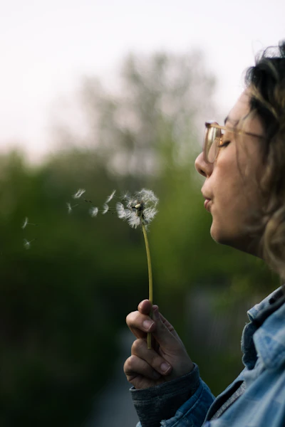 a woman blowing a dandelion with her eyes closed