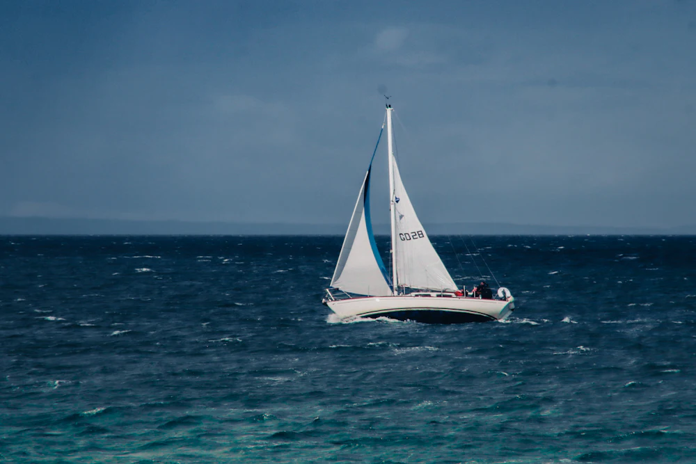 A white sloop under main and headsail crossing dark open water, whole boat in frame, low green coast behind.