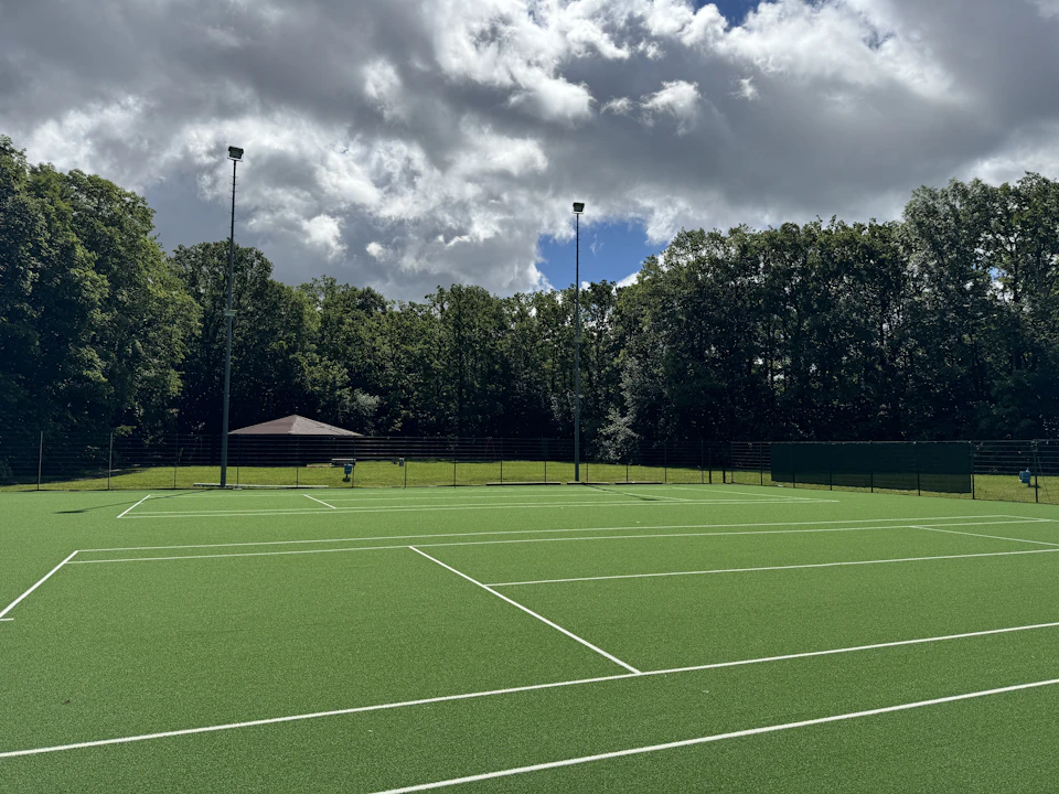 A green grass tennis court surrounded by trees under a typical English summer sky of broken clouds and sunshine, evoking Wimbledon and the British July experience