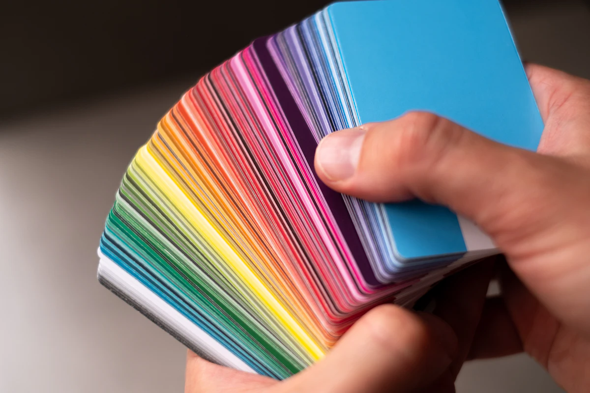 Tangan memegang kipas kartu warna yang dibuka menjadi gradasi warna pelangi, difoto dari dekat dengan latar kabur