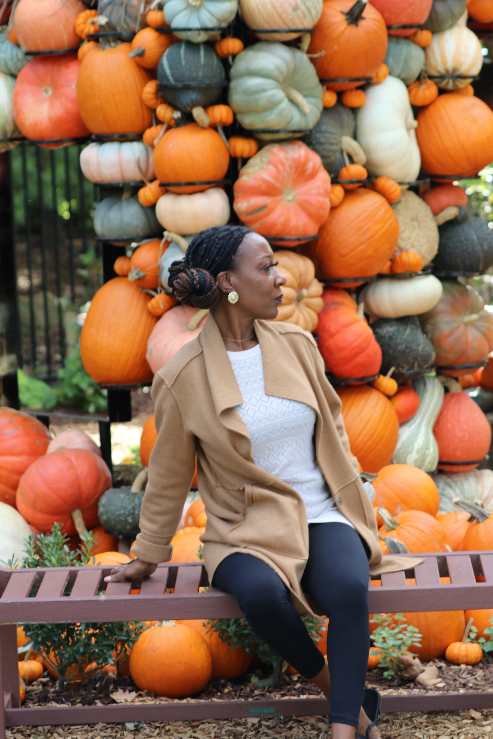 A Black woman in a camel coat sits on a bench in front of a wall of pumpkins and gourds