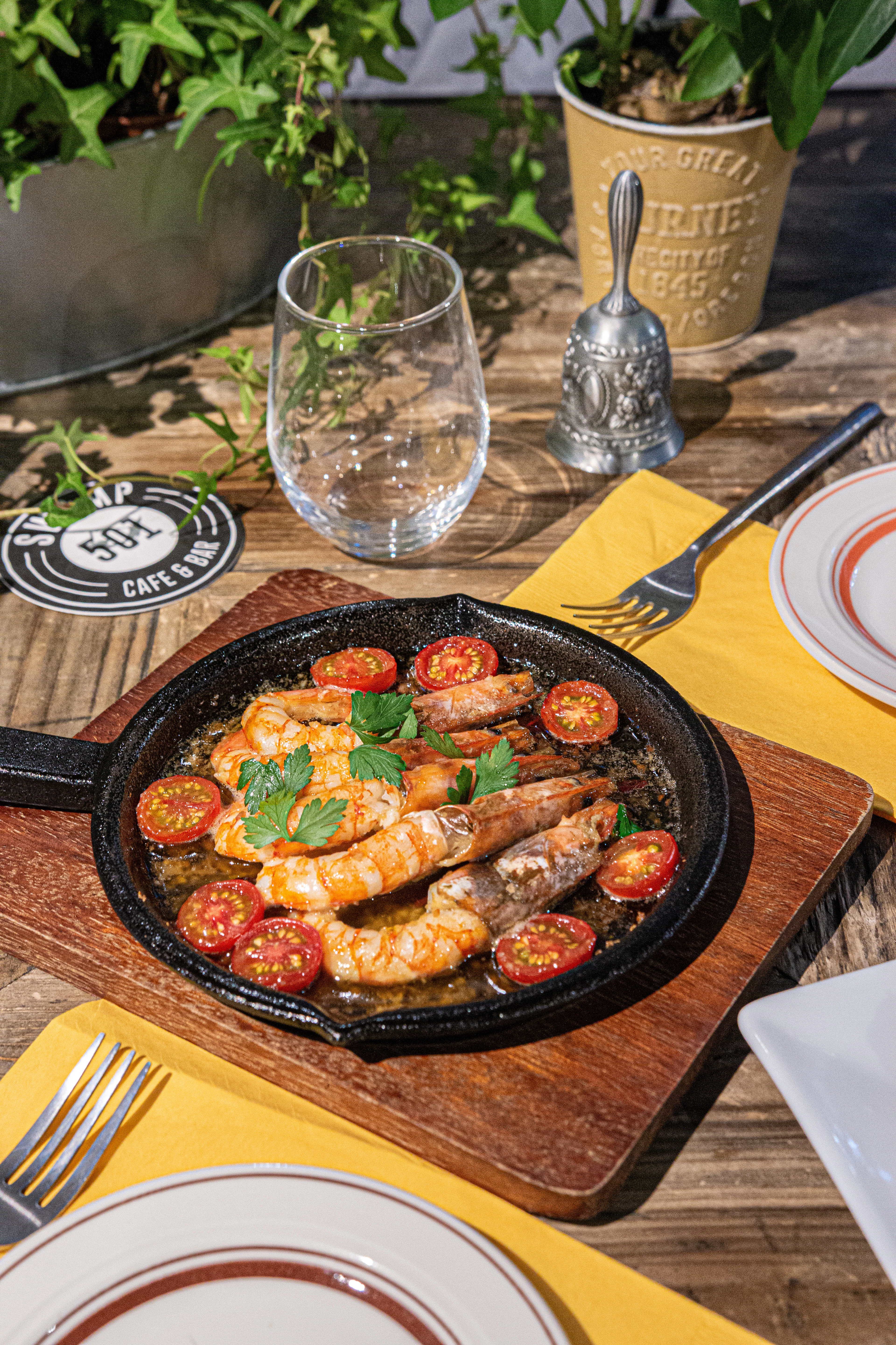A sizzling pan of prawns with tomatoes and parsley on a wooden board at a sunlit terrace table