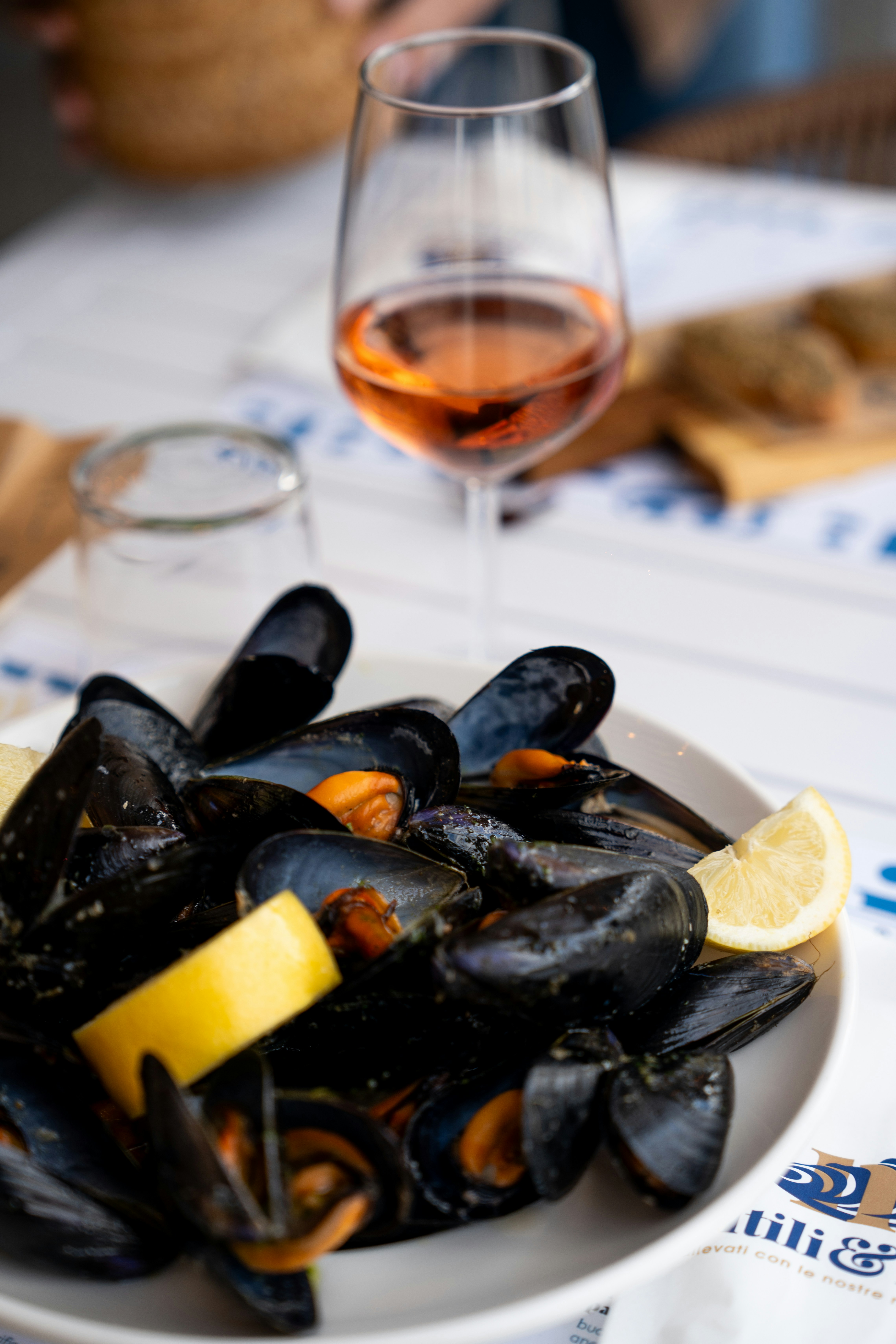 Steamed mussels with lemon wedges on a white plate, paired with rosé wine`

Image 2 is the only photo where steamed mussels are the clear, primary subject of the shot. The mussels are prominently displayed in the foreground on a plate with lemon garnish, directly relevant to the Prosecco cooking article context. The accompanying wine glass reinforces the article's theme.

Image 1: Mussels are in a wooden bowl but significantly out of focus; the wine glass in the background is sharper, making the composition ambiguous about the primary subject.

Image 3: Mussels are shown from directly above in a bowl, but the angle and presentation lack professional food photography quality for a high-quality blog.

Image 4: Mussels appear to be only partially visible in a dark pot with greens, making them not the clear primary subject