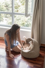 a woman kneeling down next to a cat in a cat bed