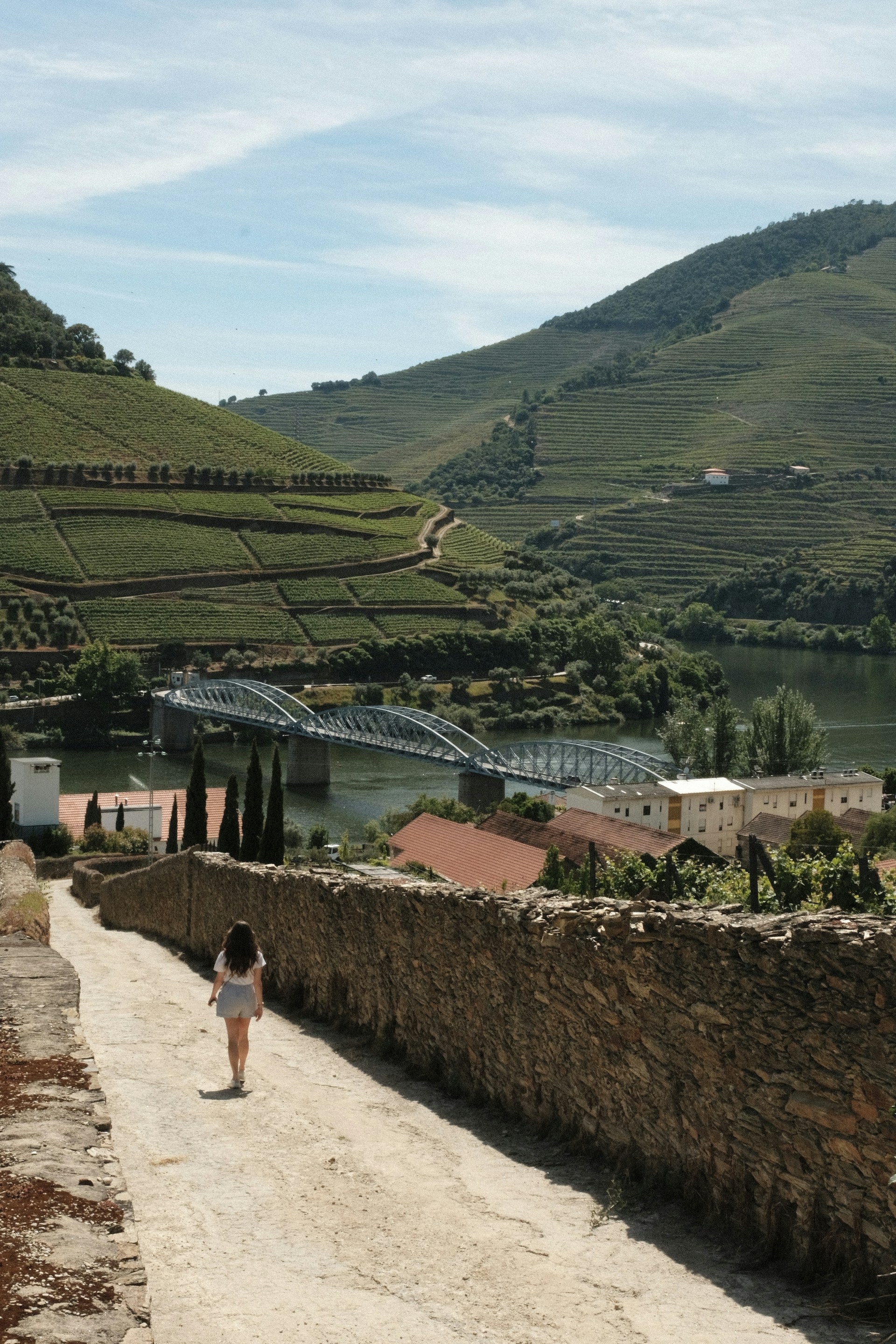 a woman walking down a dirt road next to a lush green hillside