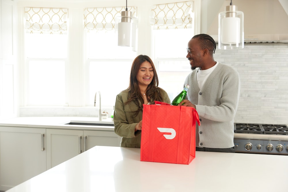 a man and woman standing in a kitchen with a red bag
