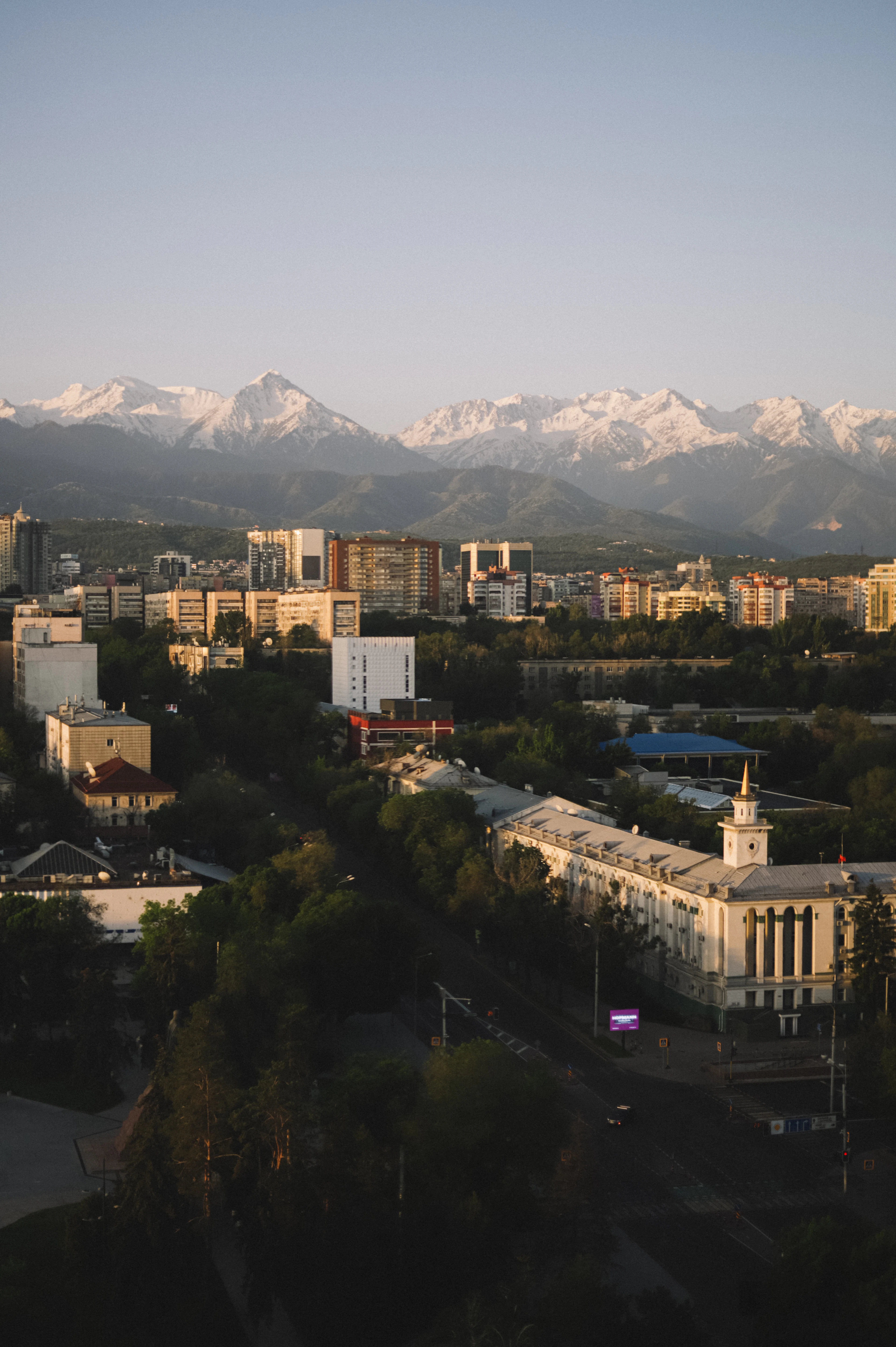 Almaty's mixed low- and high-rise skyline backed by the snow-covered Tian Shan mountains at golden hour.