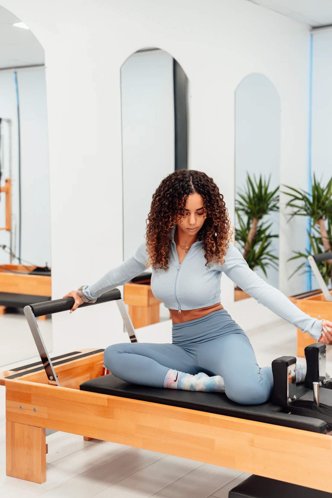 A woman in pale blue sitting tall on a wooden reformer, arms open, in a bright white studio