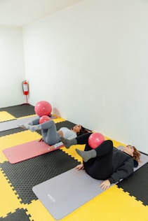 Duas mulheres deitadas no chão com uma bola pequena entre os joelhos em flexão para exercício.