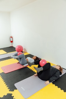 Duas mulheres deitadas no chão com uma bola pequena entre os joelhos em flexão para exercício.