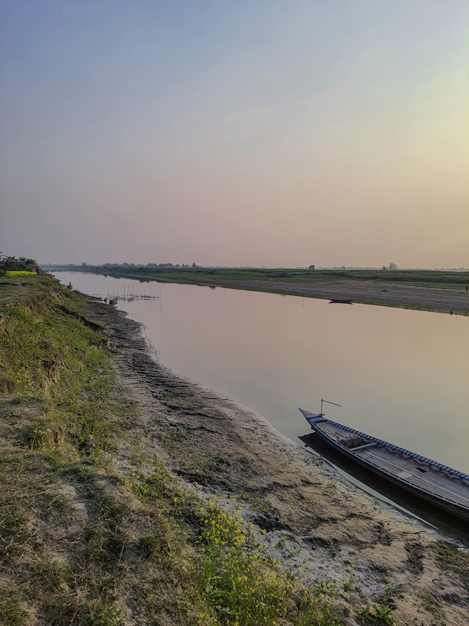 The image depicts a serene river scene with a small boat floating on its calm waters, positioned towards the right side of the frame and facing away from the camera. The riverbank is covered in lush green grass, providing a natural border to the waterway. In the distance, beyond the river bank, there are trees lining up along the horizon line, adding depth and scale to the scene. Above all this, the sky stretches out in an expanse of blue, dotted with wispy clouds that add a sense of tranquility to the image.