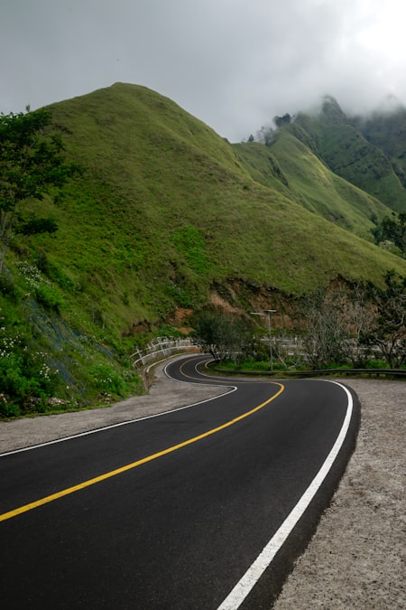 A paved road curving sharply along a green hillside with mountains in the background