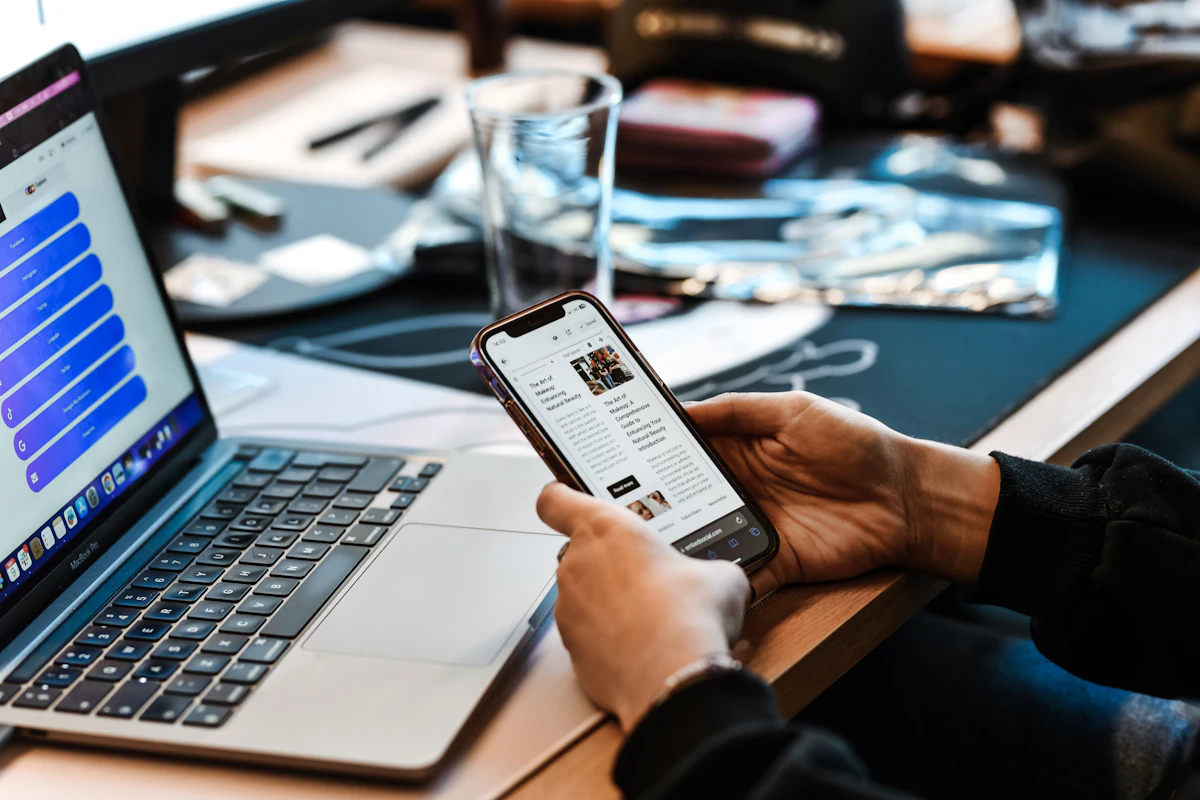 Seseorang duduk di meja dengan laptop terbuka dan ponsel di tangan, memeriksa angka mingguan