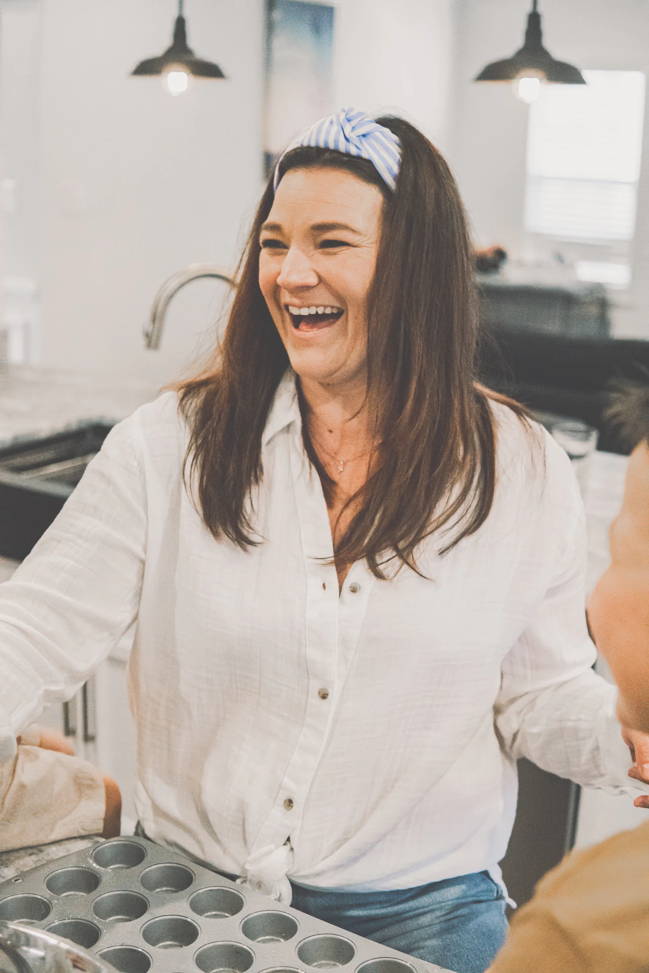 Laughing baker in a white shirt and headband holds a muffin tray in a bright kitchen.