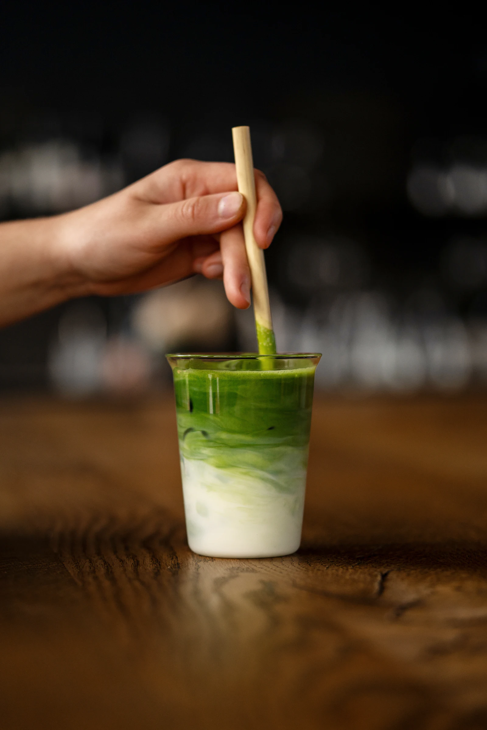A hand stirring a small glass of layered iced matcha latte on a wooden bar