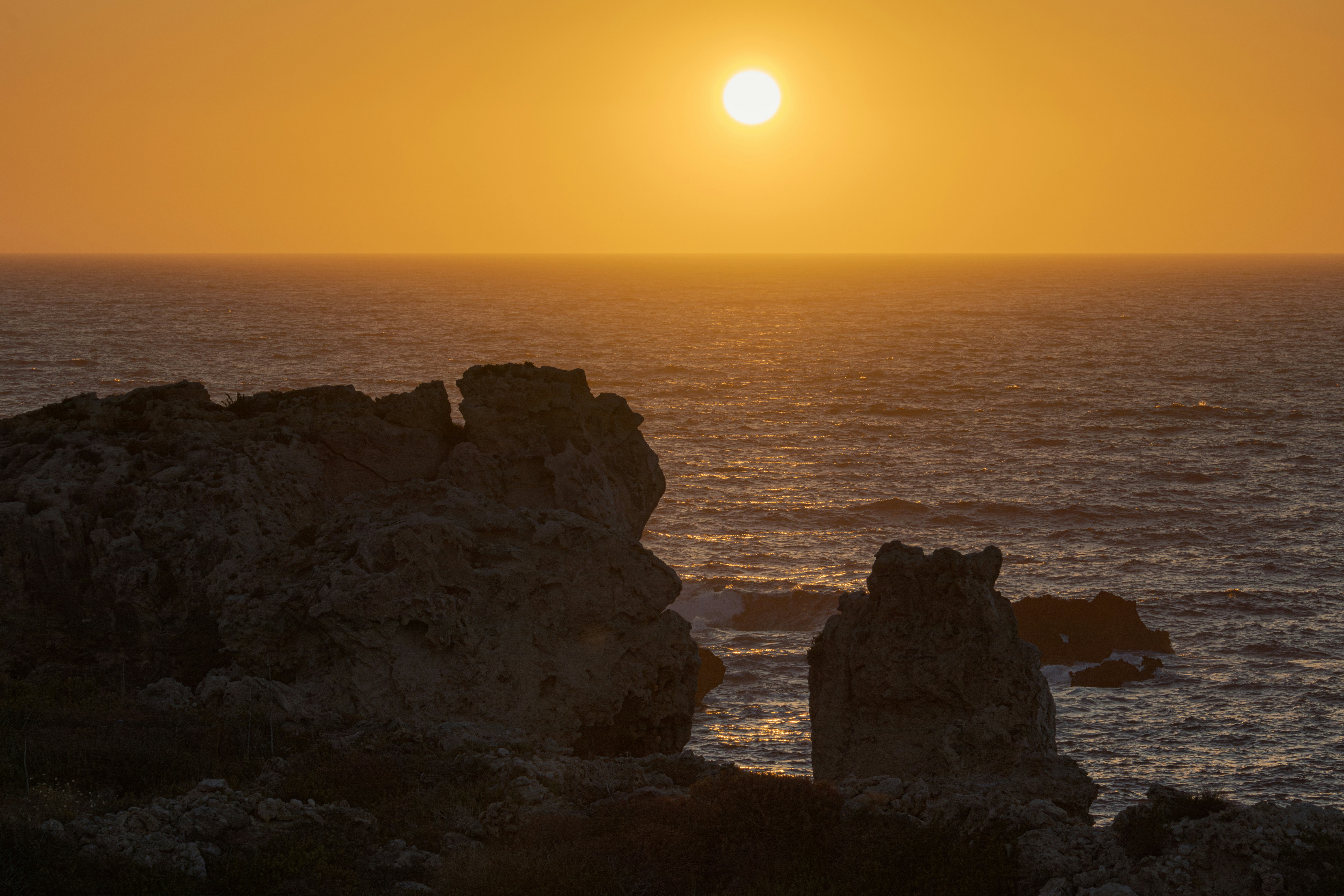 the sun is setting over the ocean with rocks in the foreground, 