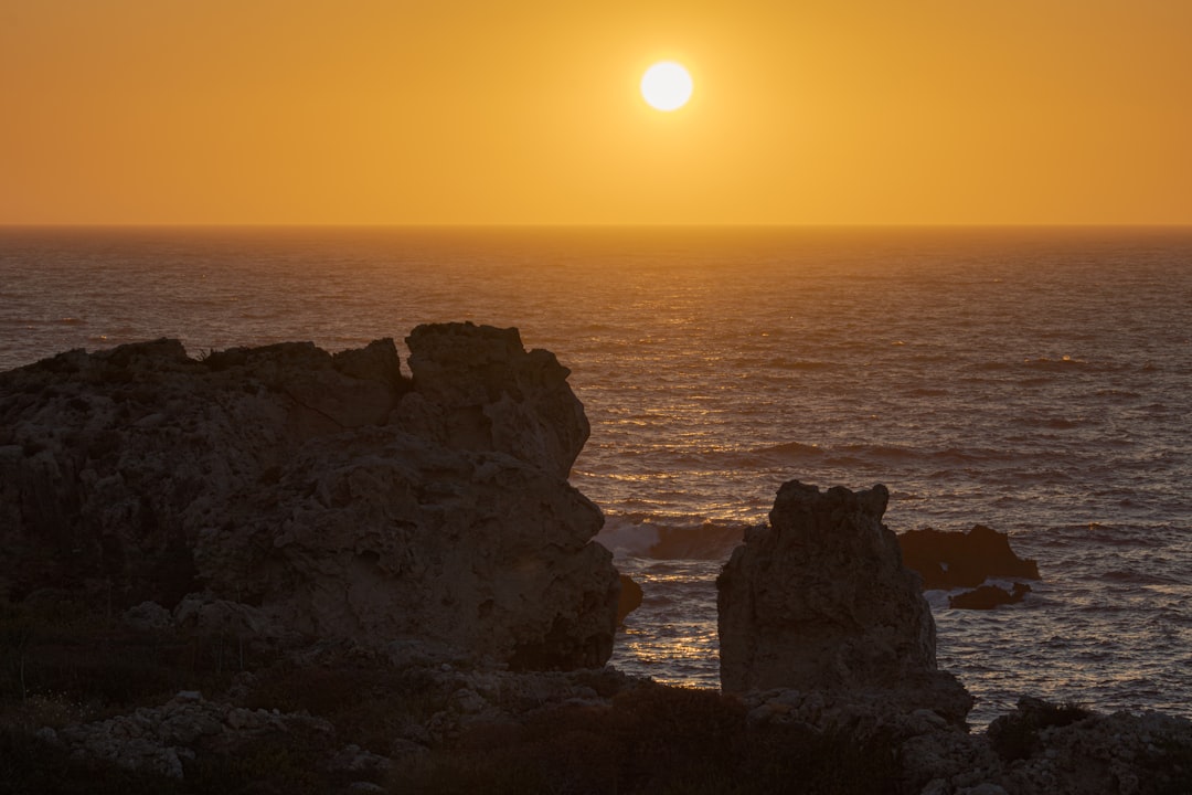 the sun is setting over the ocean with rocks in the foreground,