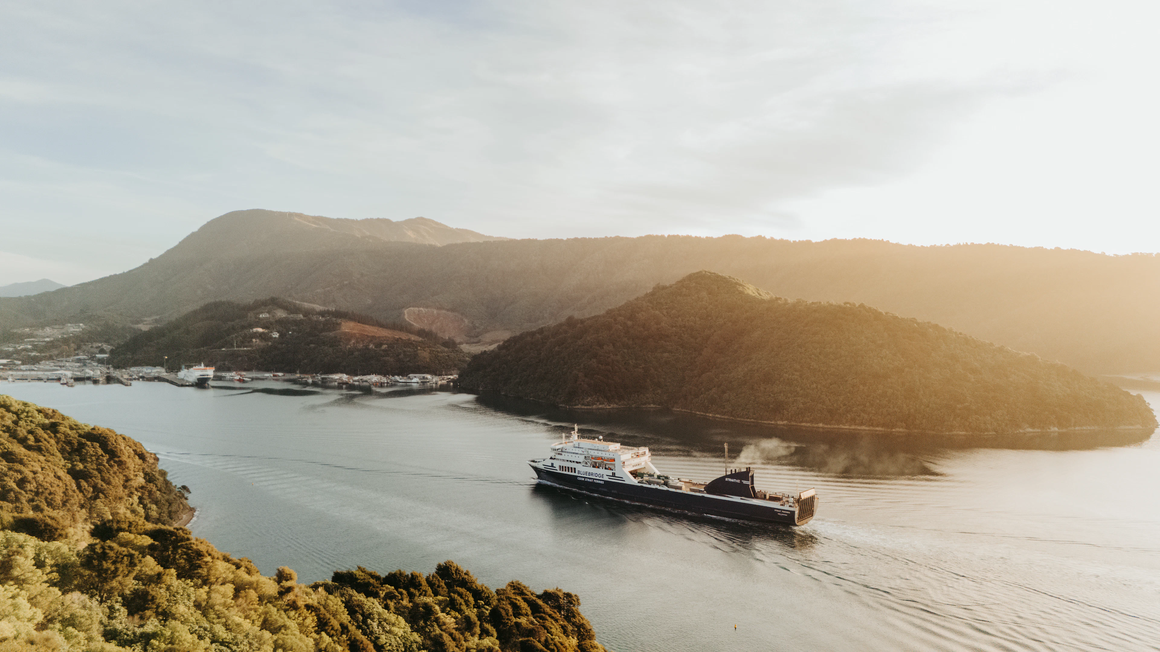 A ferry crossing calm water between forested headlands in low golden light, New Zealand
