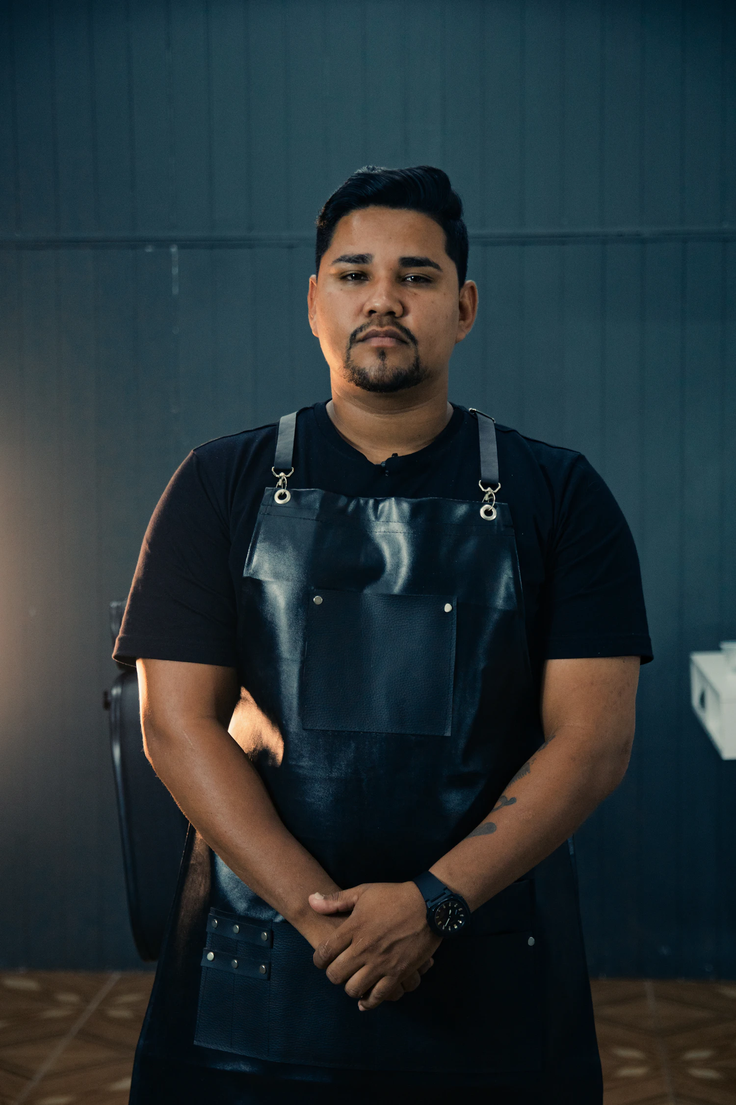 A barber in a black apron photographed straight on against a dark wall
