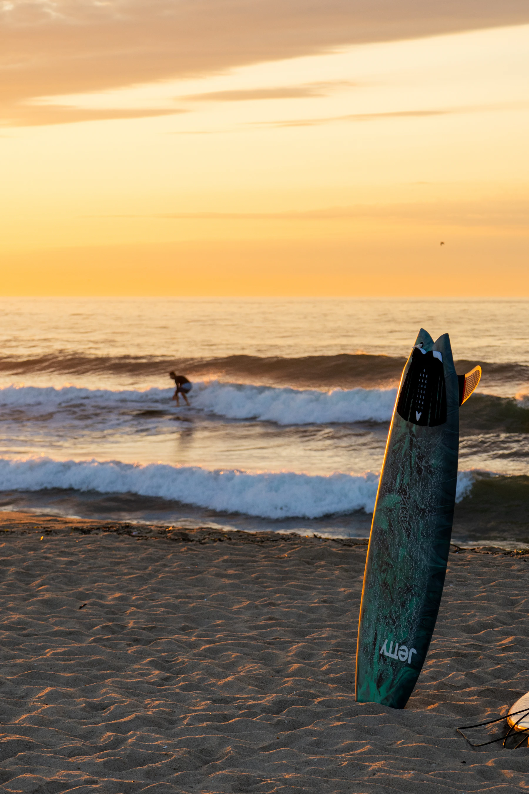 A surfboard standing in the sand at dawn, a surfer on the wave behind
