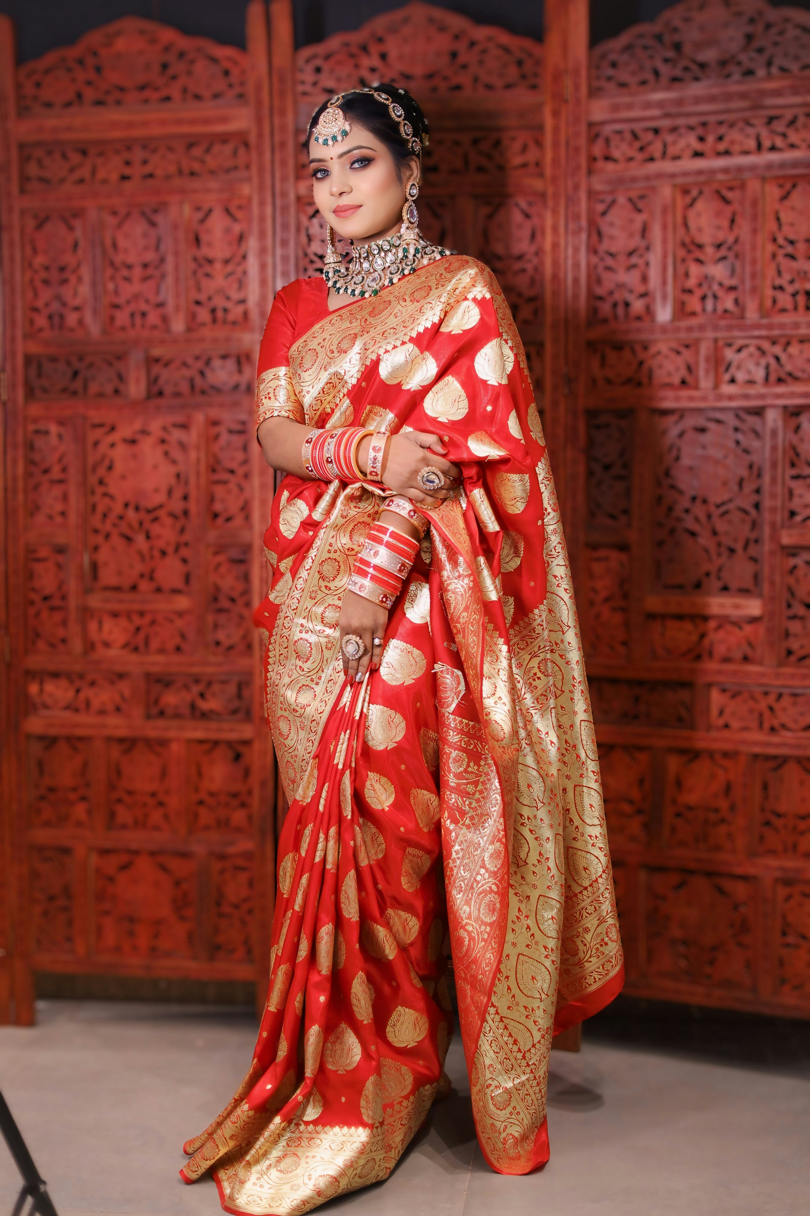 A woman in a red and gold saree stands before carved wood, adorned with jewelry, radiating festive elegance and tradition.