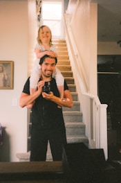 a man holding a little girl on his shoulders in a family home with stairs