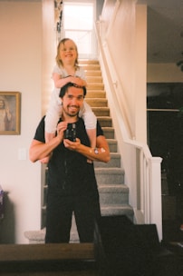 a man holding a little girl on his shoulders in a family home with stairs