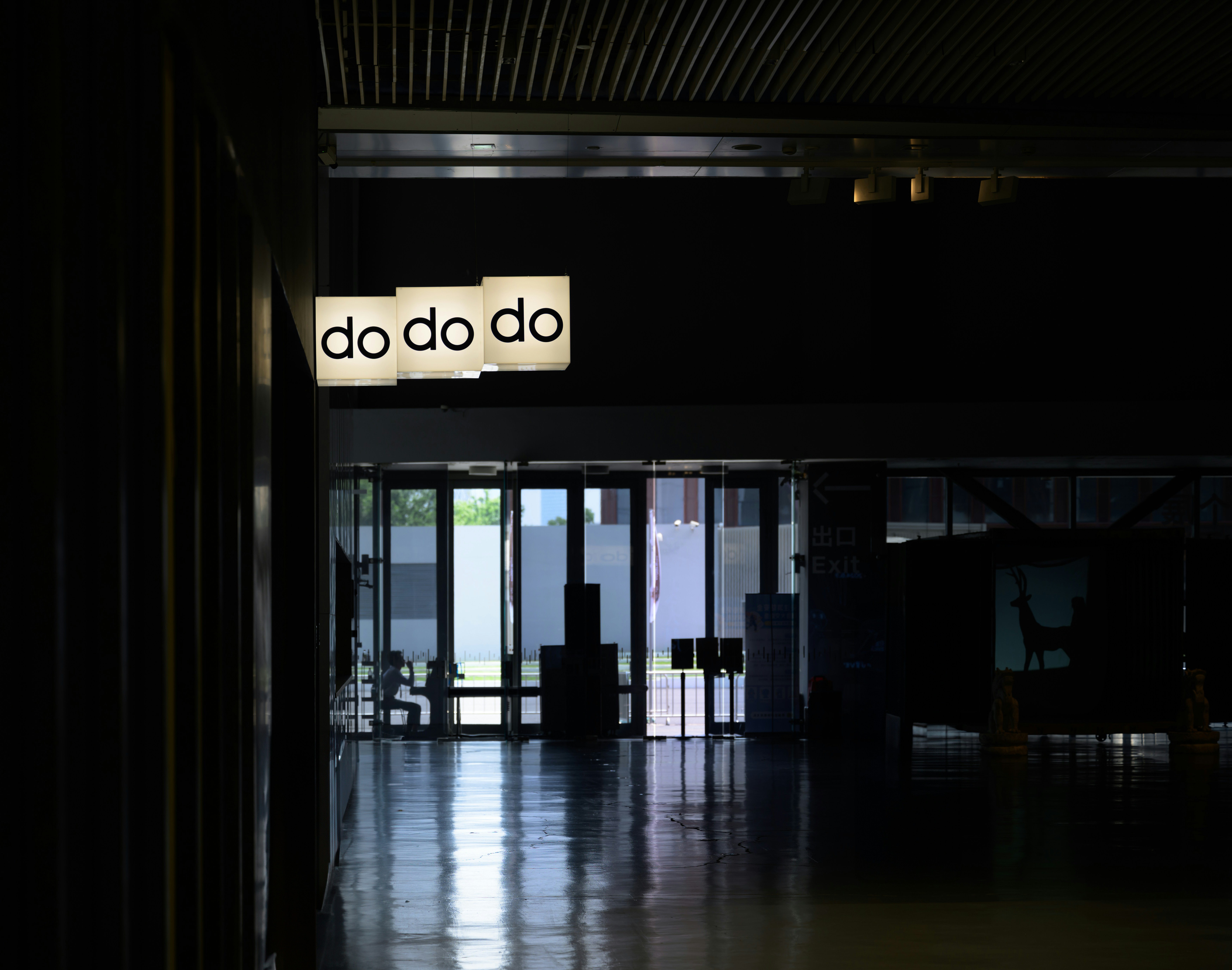 At the entrance of a venue, there was a person sitting there, and there was a light box advertisement that said do do do.