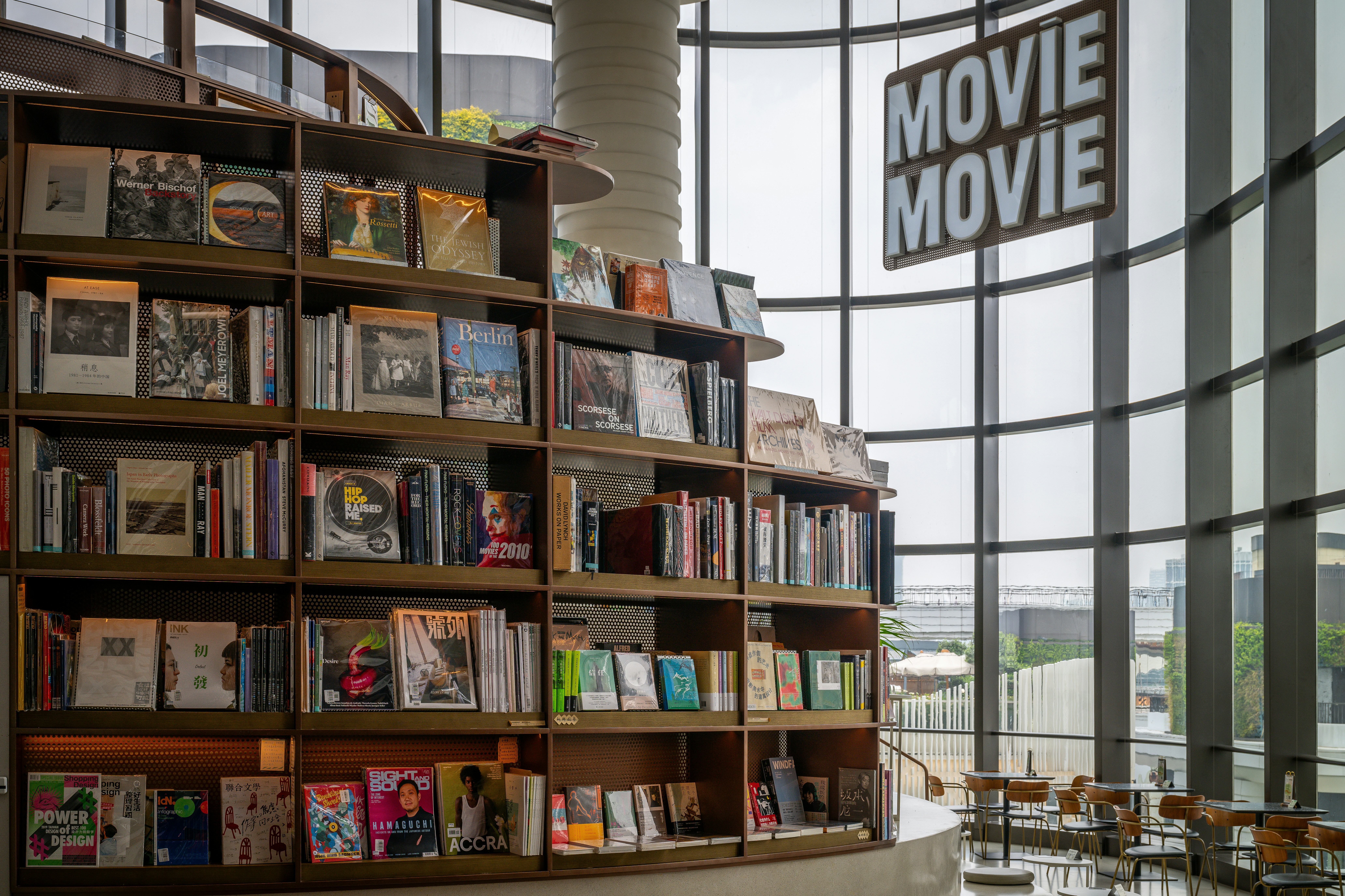 In the corner with the floor-to-ceiling windows, there is a shelf with many books and some tables and chairs around it.