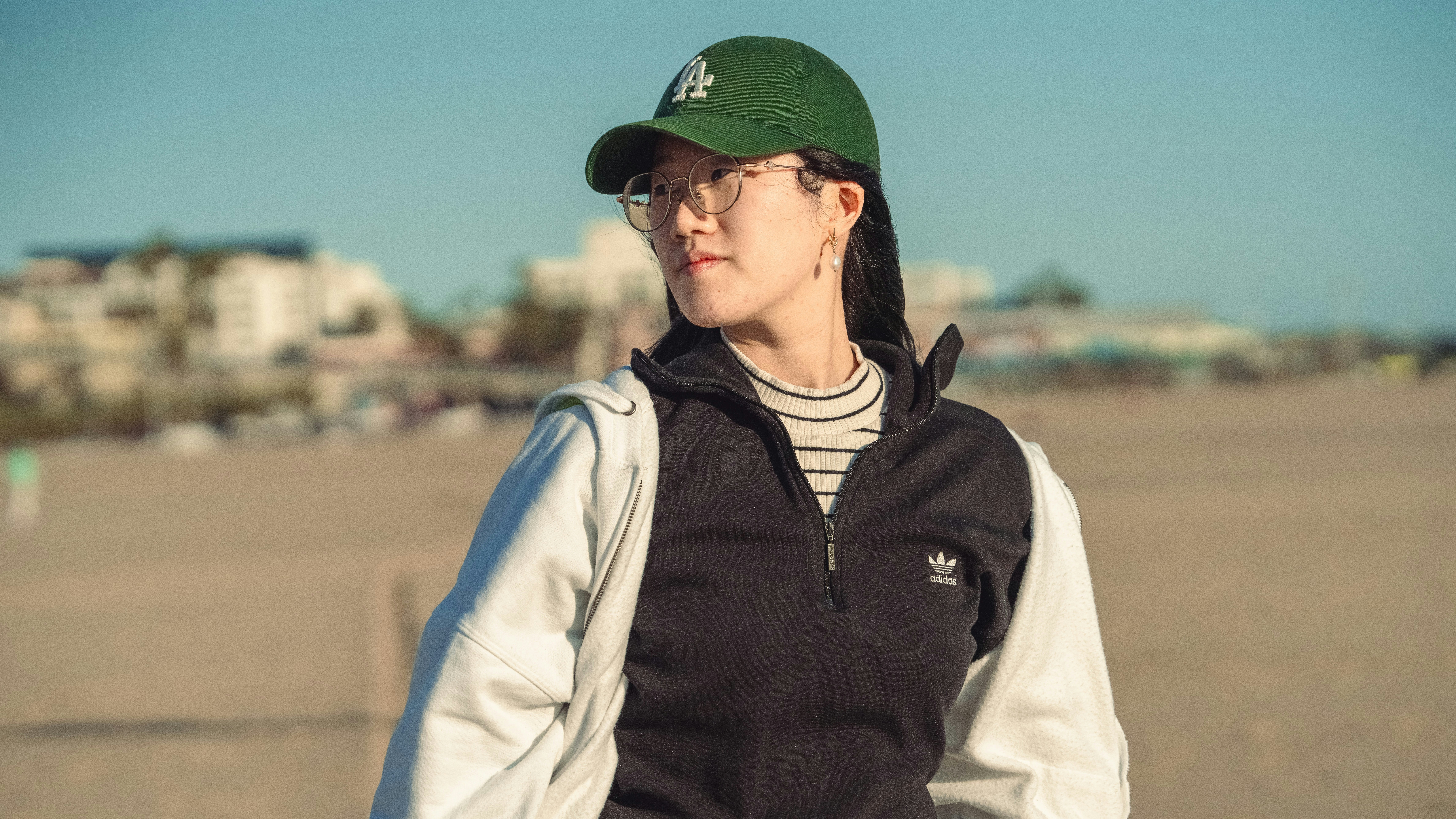 Golden hour on Santa Monica Beach: she stands relaxed in a green LA cap and round glasses, sunlight catching the edge of her striped tee beneath a black quarter-zip. The blurred backdrop of beachfront apartments and pale sand sets a laid-back, urban-coastal vibe as she glances off-frame, looking effortlessly cool in the warm seaside breeze.