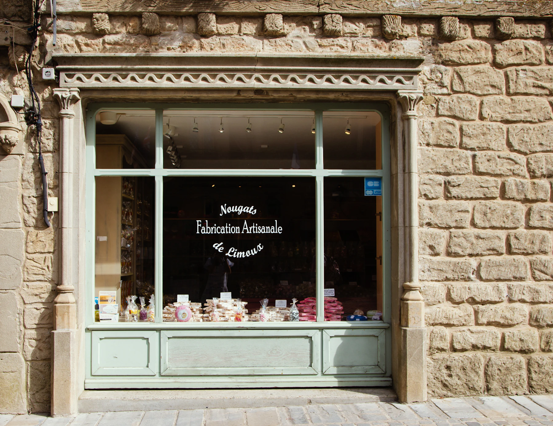 A pale green shop window set in a stone wall in France, lettered in white French script, with sweets on display